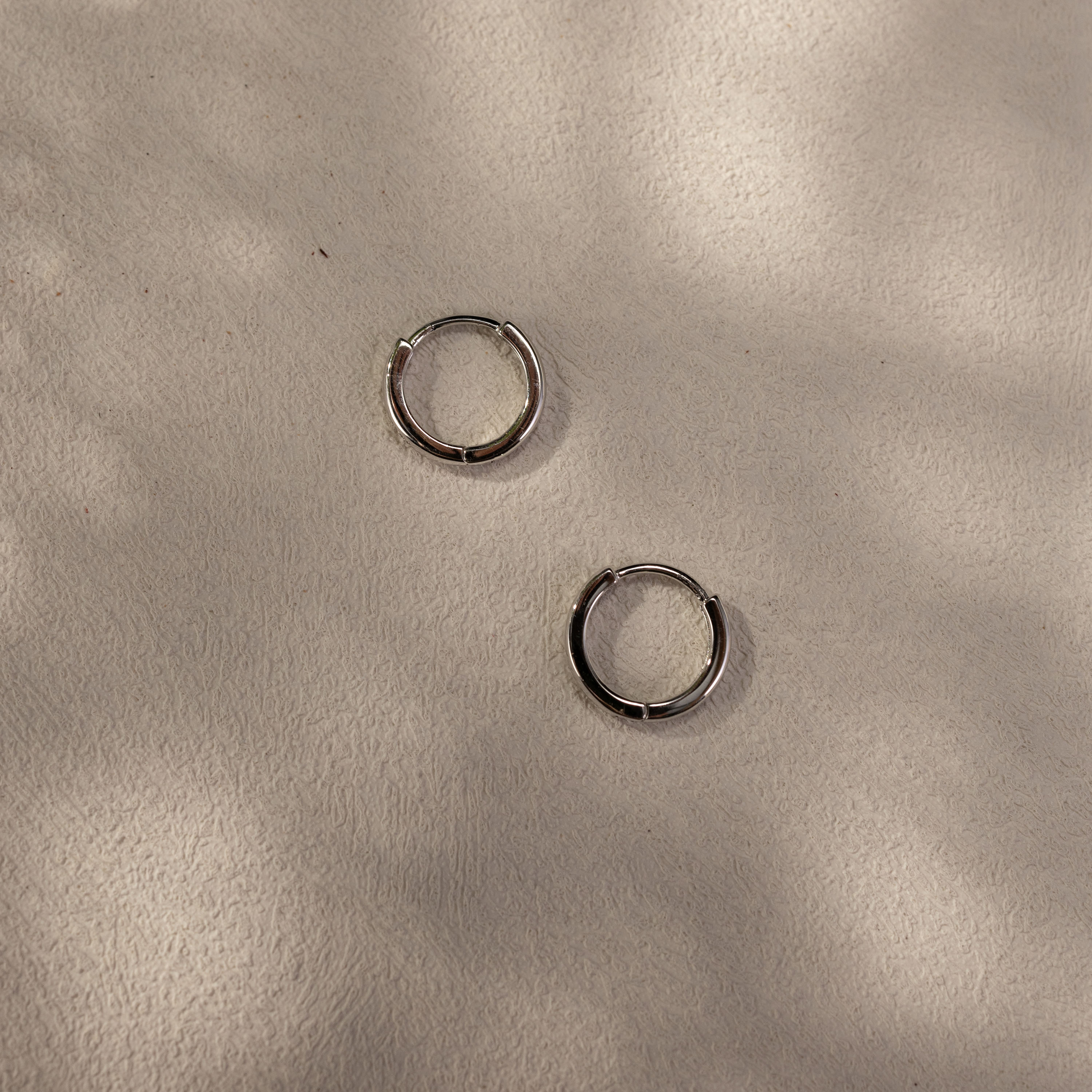 Minimalist silver Lina huggie earrings placed on a soft textured surface with natural soft shadows, lightweight waterproof everyday hoops by SILVBREEZE