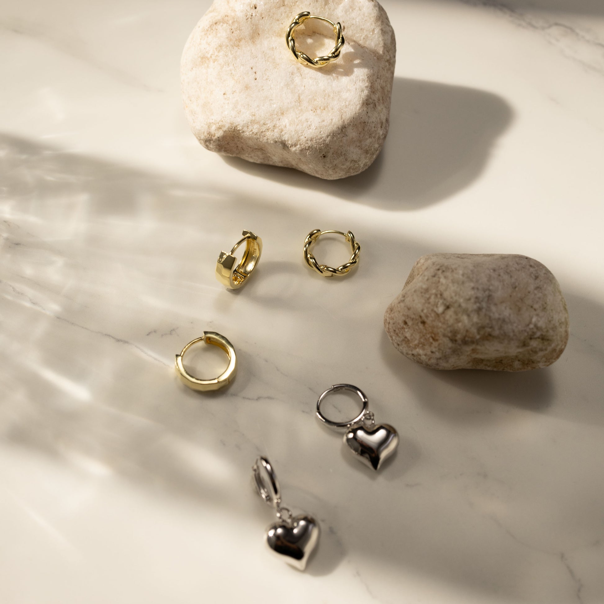 Flatlay of Mira gold huggie earrings styled with other twisted gold and silver heart huggies on a light stone-textured surface, showcasing a mixed athleisure earring stack by SILVBREEZE.