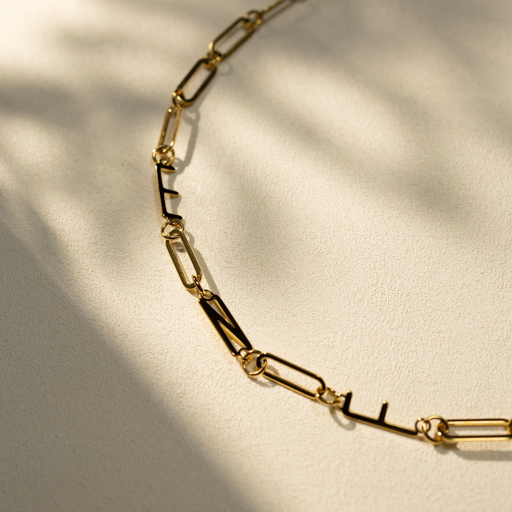 Closeup of a gold MBTI letter necklace on a warm textured surface, featuring paperclip chain links and natural shadow details. Minimal lifestyle product shot.