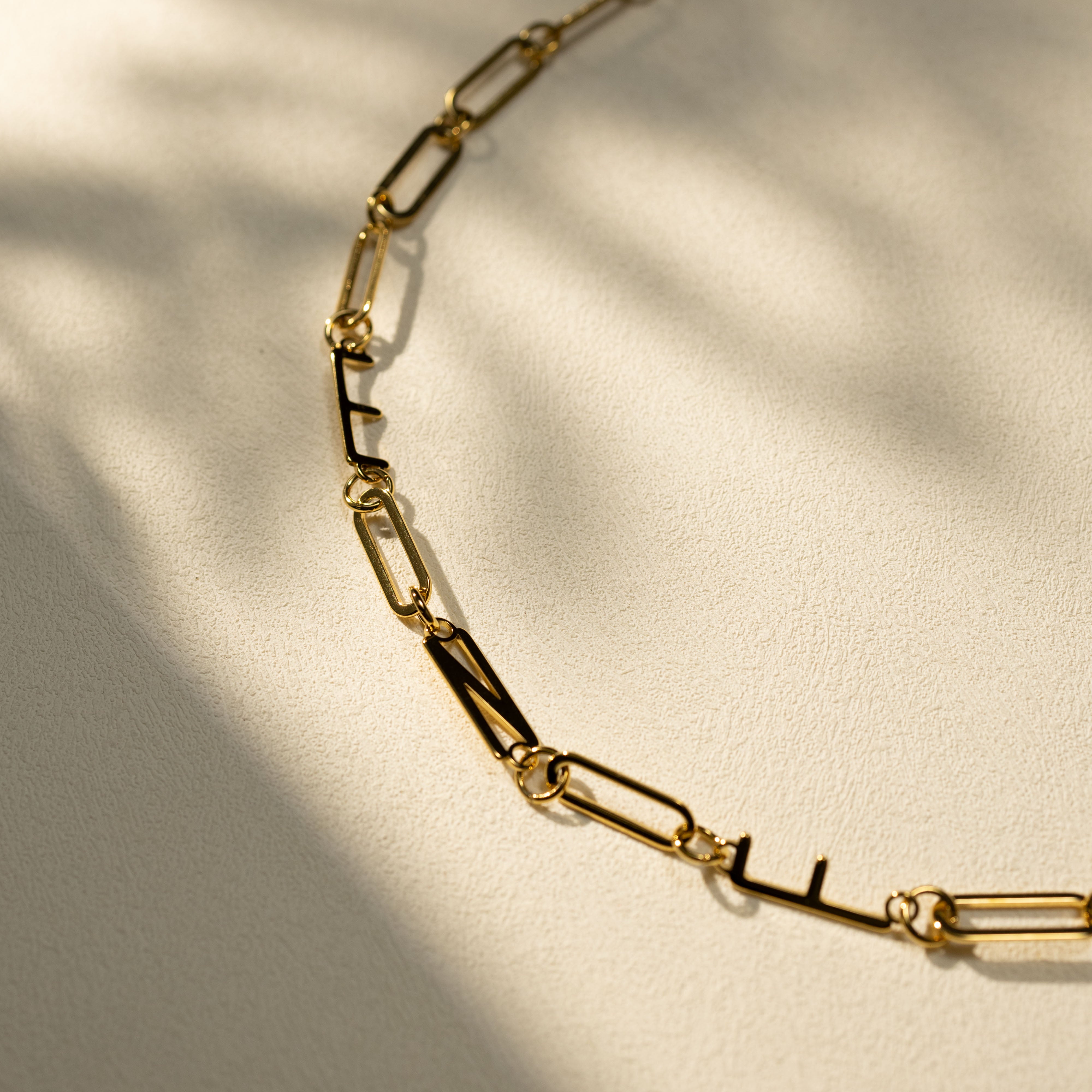 Closeup of a gold MBTI letter necklace on a warm textured surface, featuring paperclip chain links and natural shadow details. Minimal lifestyle product shot.