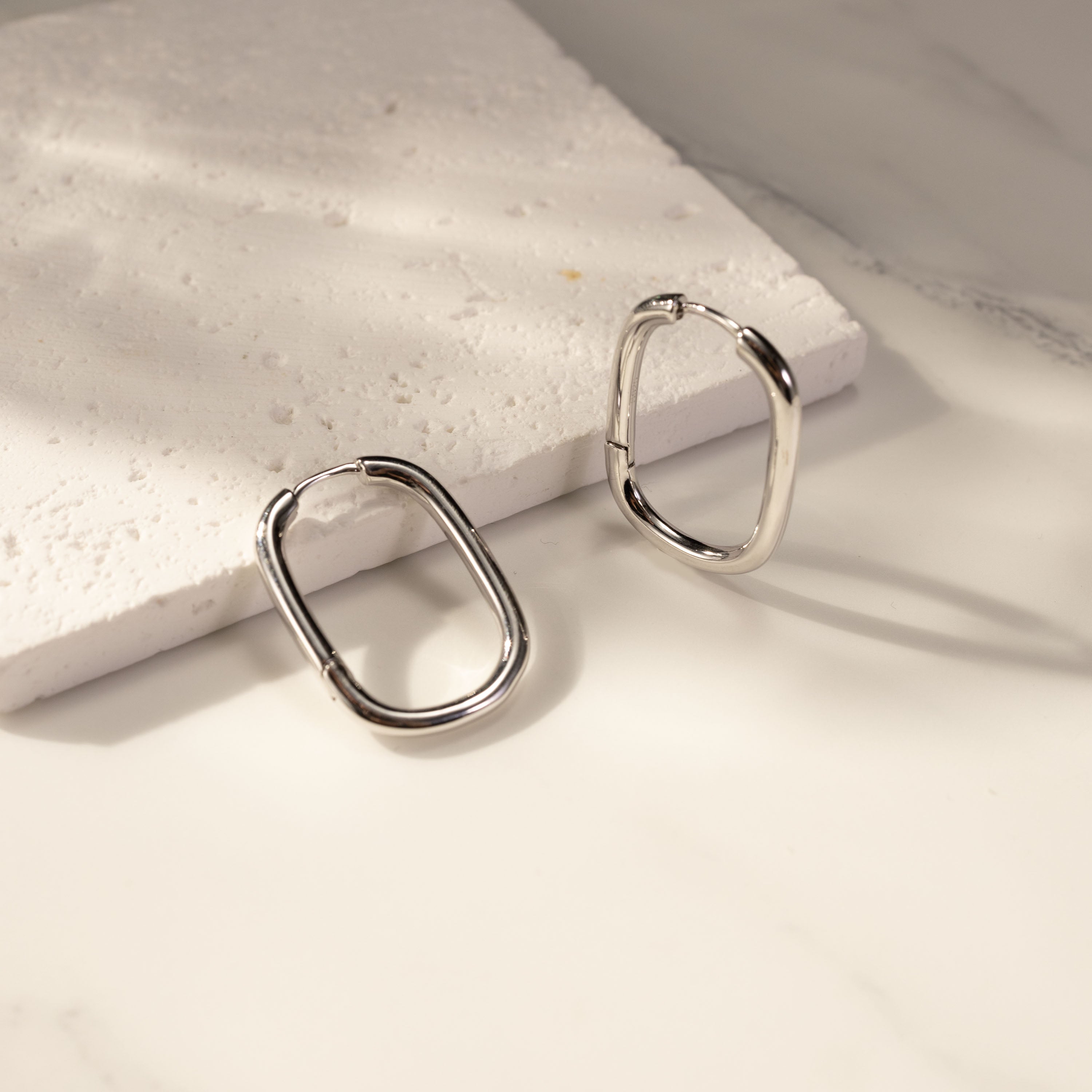 Silvbreeze Athena silver hoop earrings displayed on a textured white stone surface, showcasing the sleek oval silhouette and polished shine.