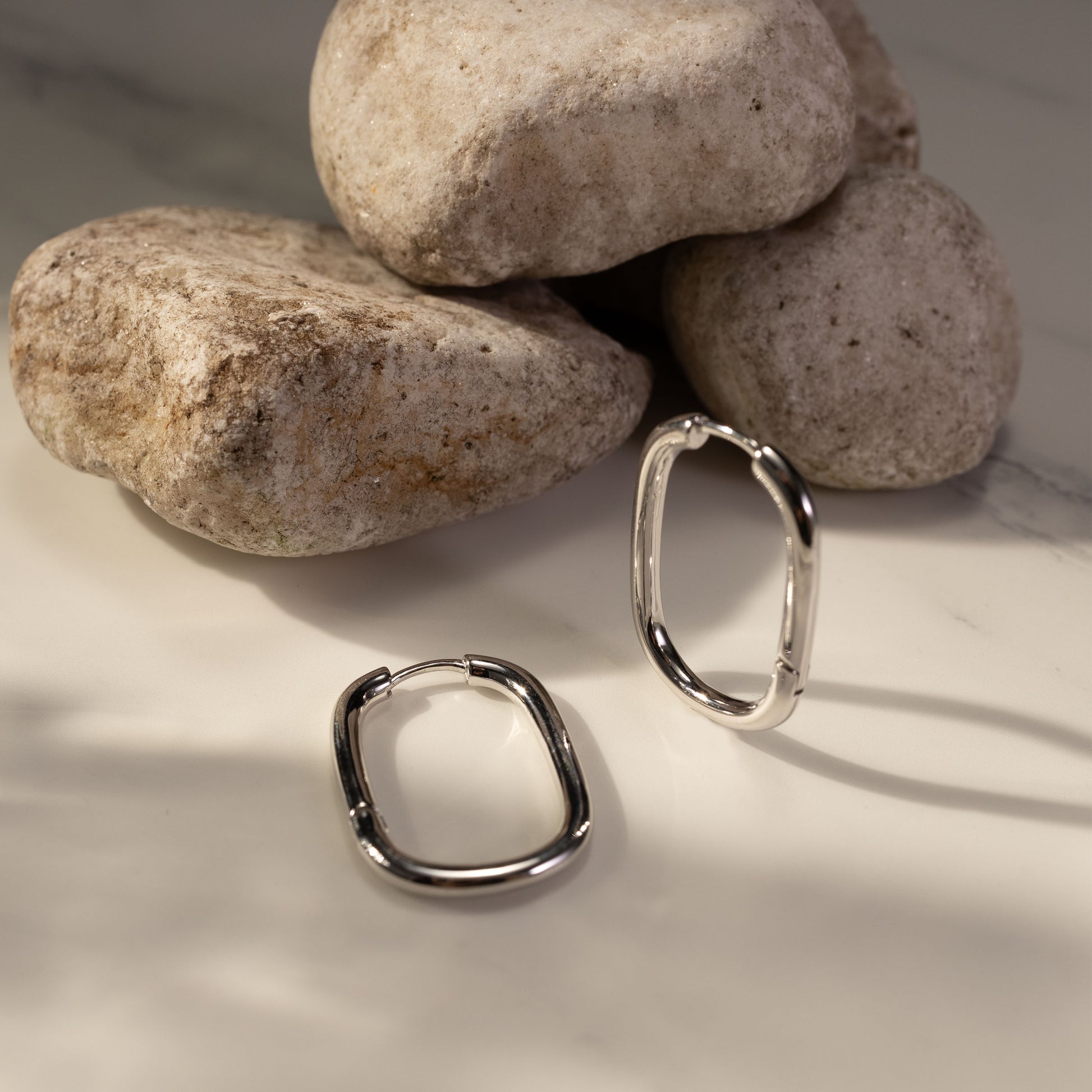 Silvbreeze Athena silver hoop earrings styled on smooth natural stones, highlighting the polished oval design and minimalist shine.