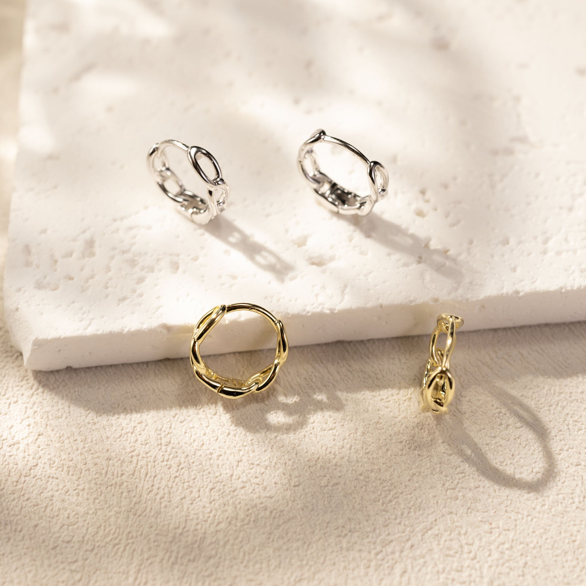 Eva twisted link huggie hoop earrings by SILVBREEZE in white gold vermeil and 14K gold vermeil, crafted from solid sterling silver, displayed together on a textured stone surface with warm sunlight and soft shadows in a minimalist lifestyle jewelry scene