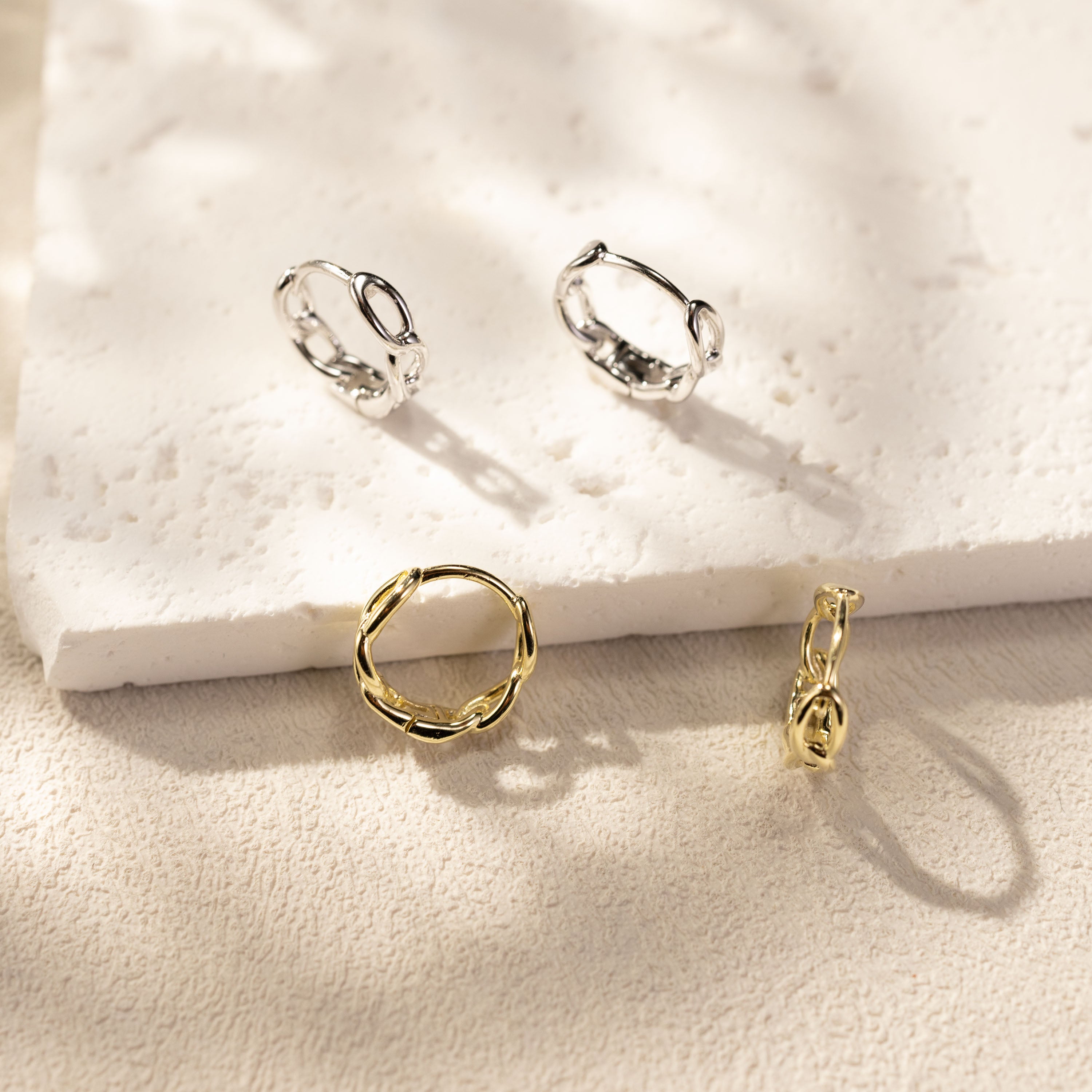 Eva twisted link huggie hoop earrings by SILVBREEZE in white gold vermeil and 14K gold vermeil, crafted from solid sterling silver, displayed together on a textured stone surface with warm sunlight and soft shadows in a minimalist lifestyle jewelry scene