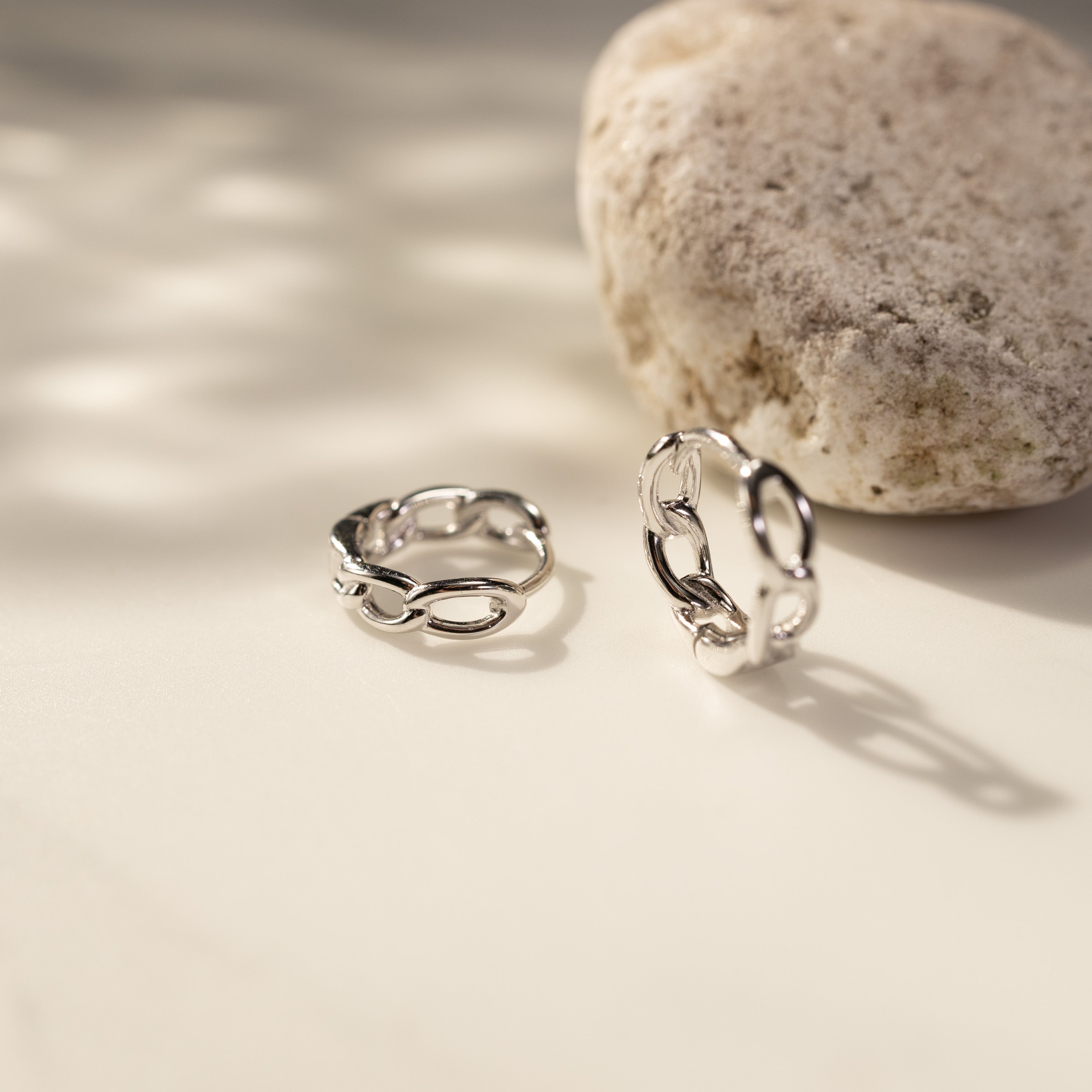 Eva white gold vermeil twisted link huggie hoop earrings made from solid sterling silver, displayed on a clean neutral surface beside a natural stone, with soft sunlight and organic shadow patterns creating an elegant minimalist lifestyle jewelry scene