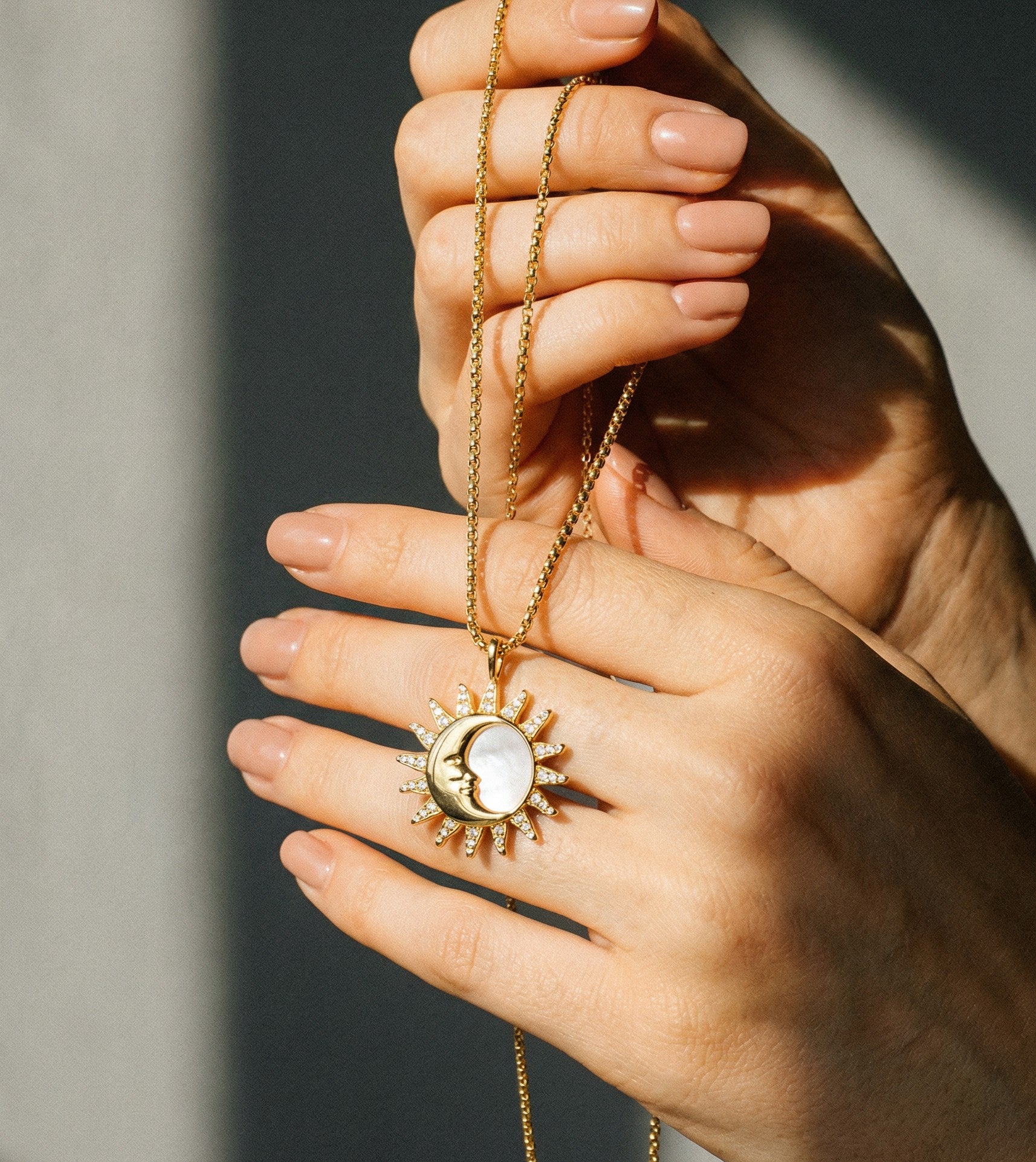 A close-up hand held shot of the Silvbreeze gold sun and moon necklace, showing the intricate details of the mother of pearl inlay and crystal-paved rays on a round box chain under natural light.