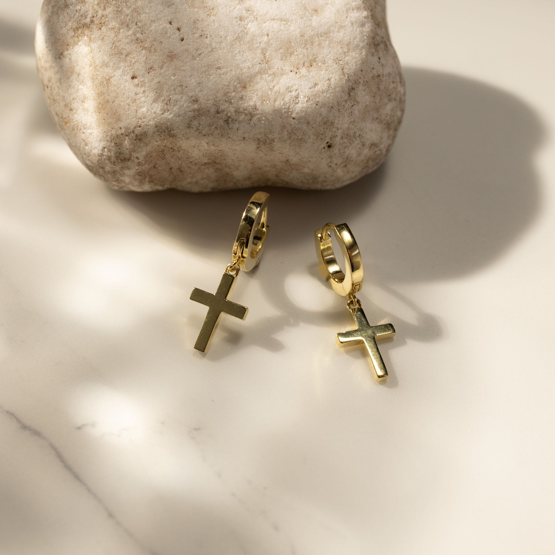 Silvbreeze Halo gold vermeil cross huggie earrings displayed in soft lifestyle scene with natural light and stone prop, minimalist modern dangling cross hoop jewelry