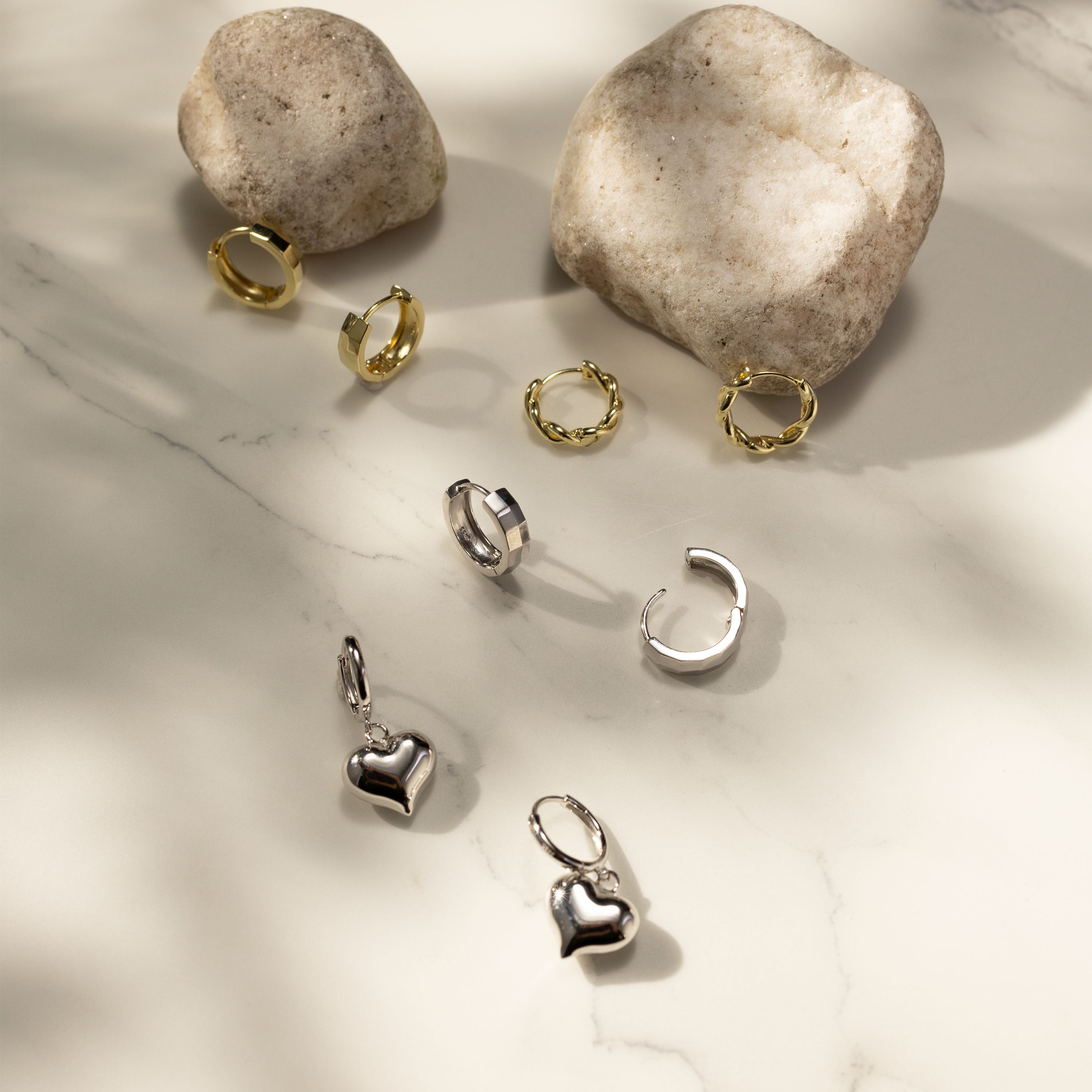 Flatlay of Silvbreeze 14K gold vermeil huggies and 925 silver heart drop earrings styled on a light stone surface.