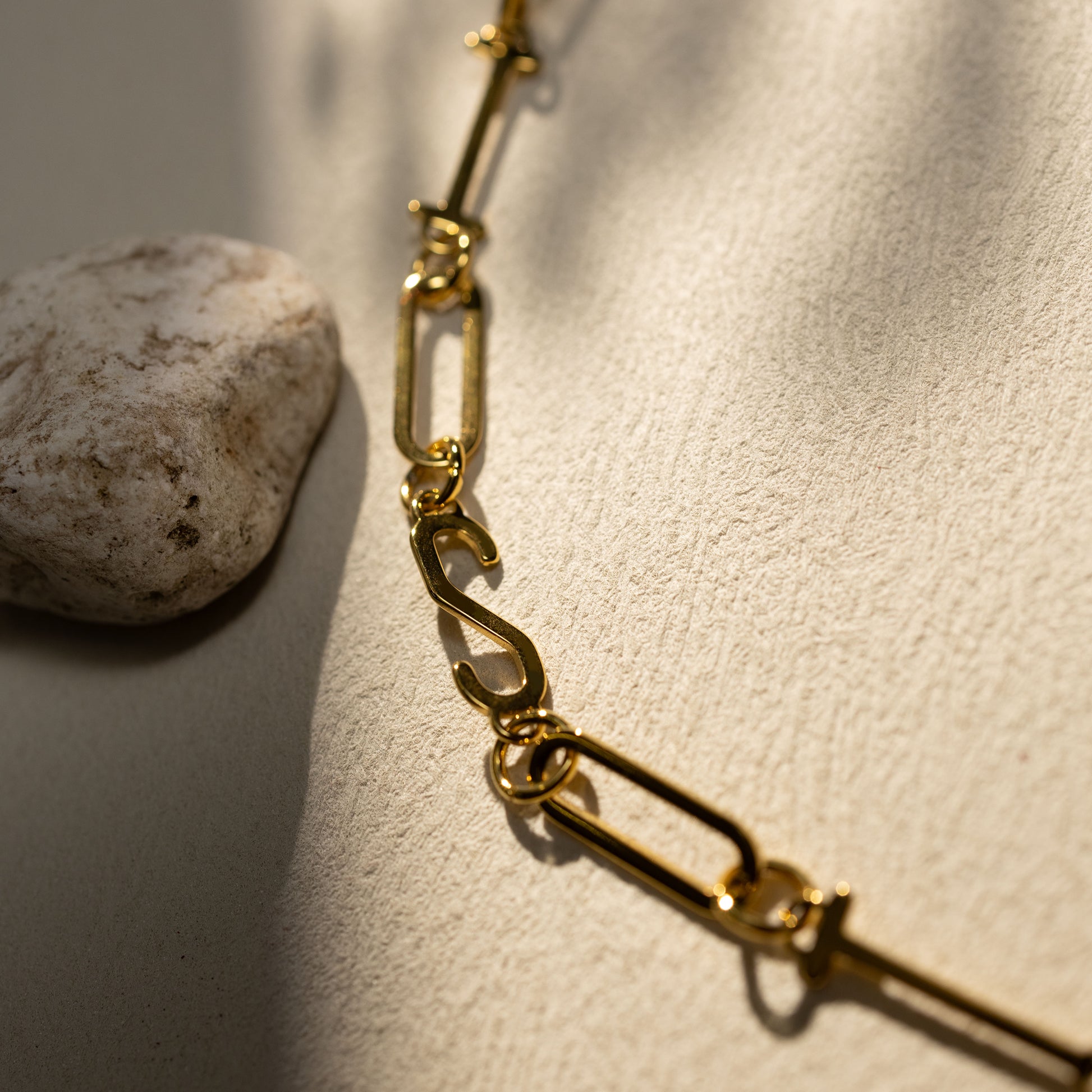 Closeup of the Silvbreeze ISTP 18K gold paperclip chain necklace featuring the letter “S”, shown on a warm neutral textured surface with soft natural shadows.