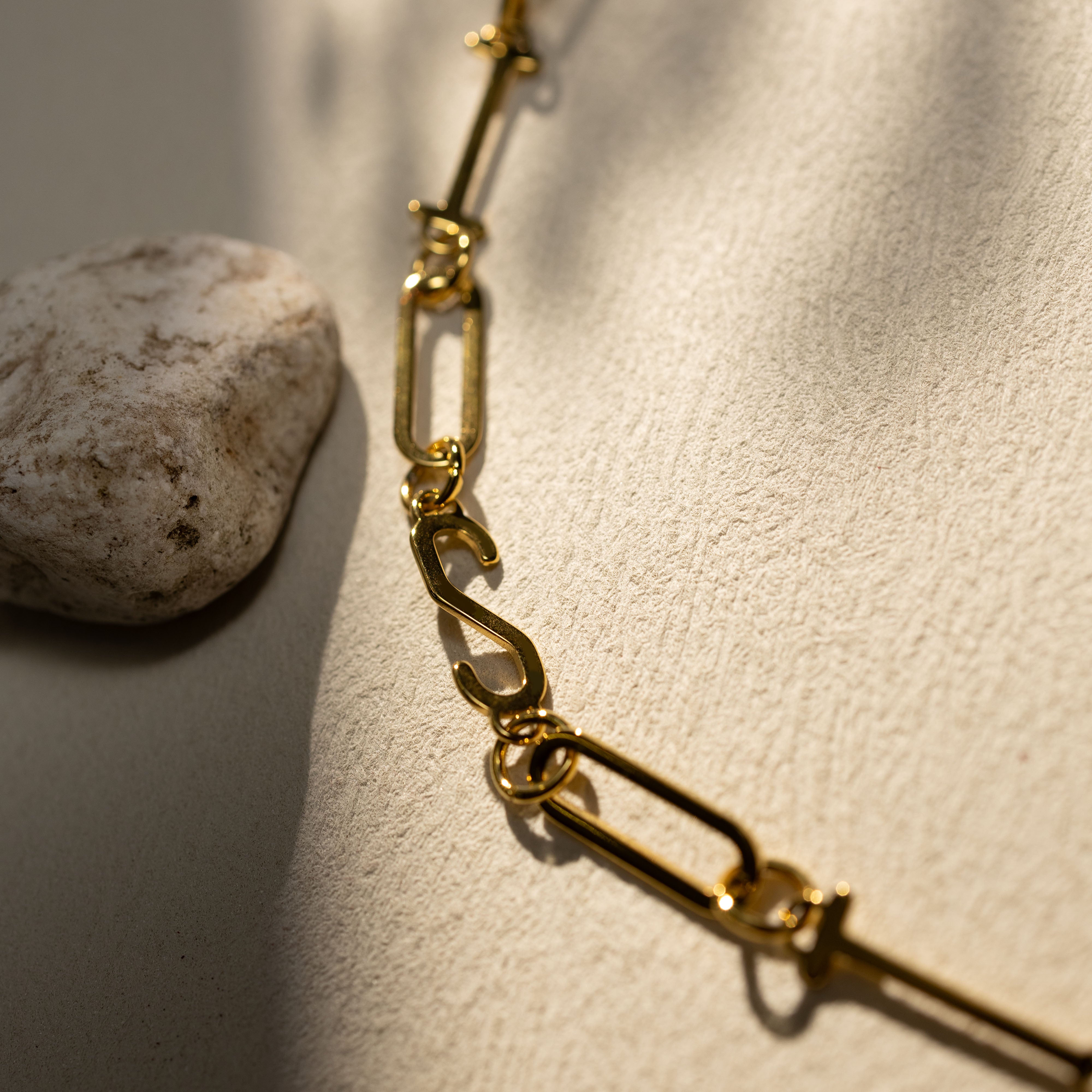 Closeup of the Silvbreeze ISTP 18K gold paperclip chain necklace featuring the letter “S”, shown on a warm neutral textured surface with soft natural shadows.