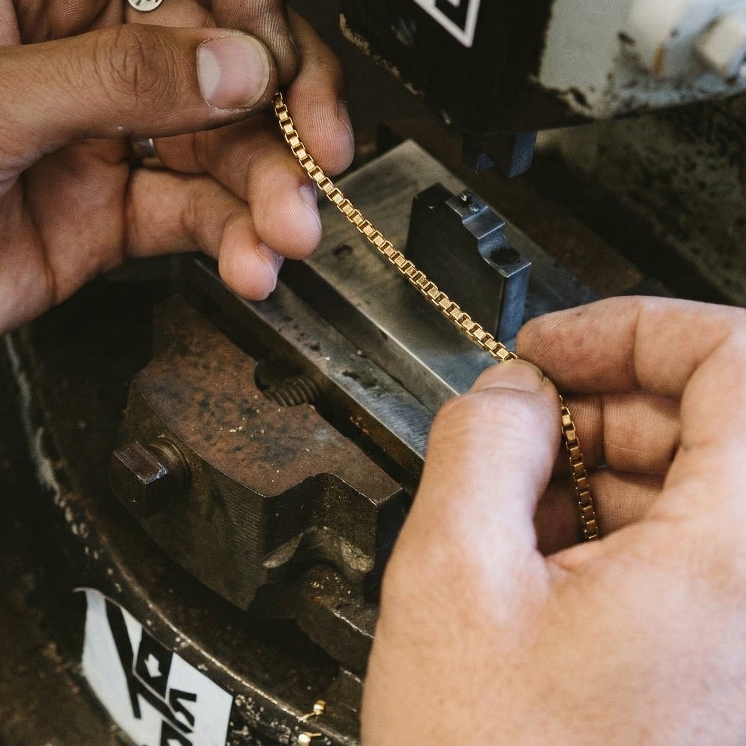 Jewelry craftsman hand polishing a gold box chain in a workshop, close-up of hands and metal tools, showcasing craftsmanship and detail.