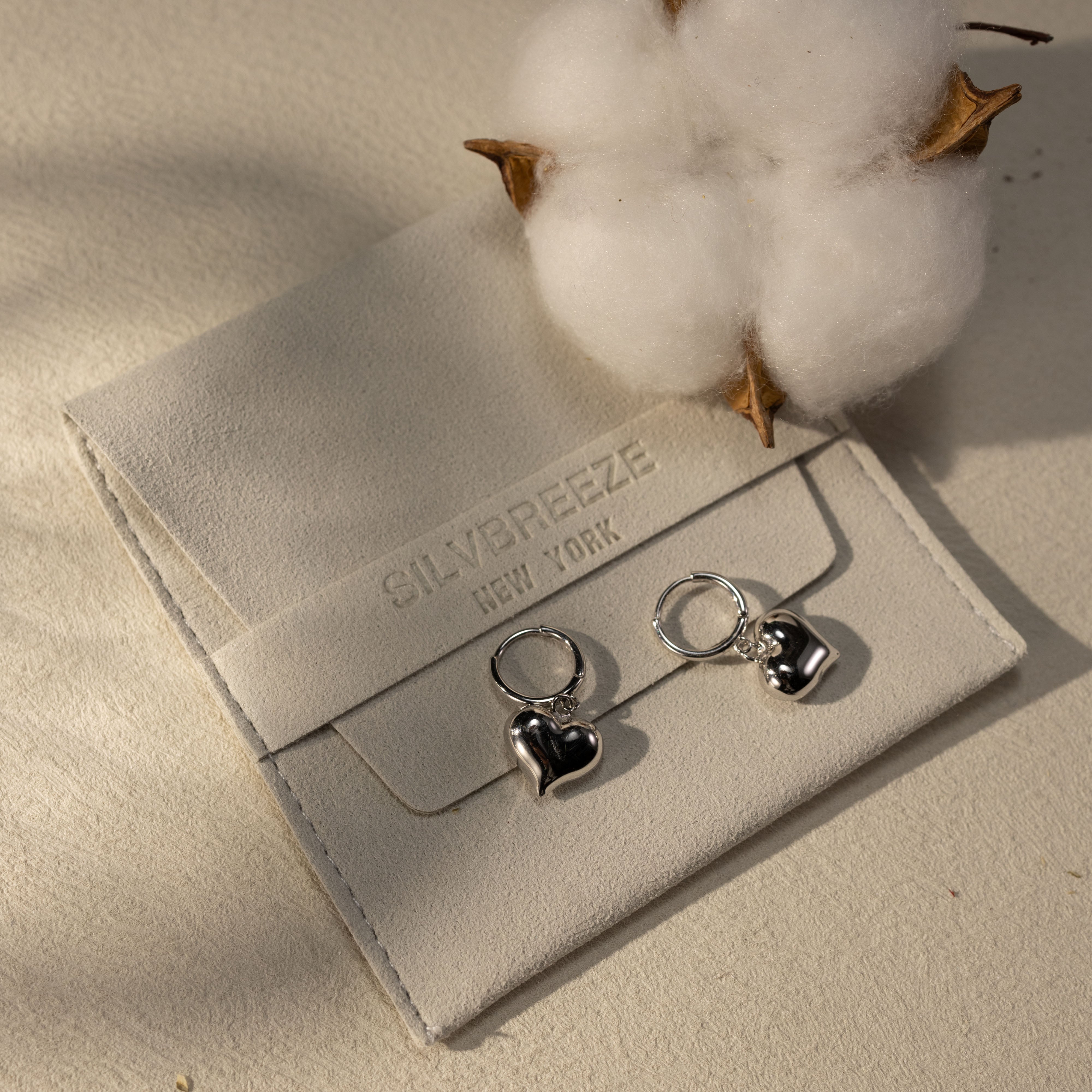 Silvbreeze Lune white gold vermeil heart dangle huggie earrings displayed on soft suede jewelry pouch with cotton detail, sterling silver waterproof hypoallergenic earrings in a warm lifestyle setting