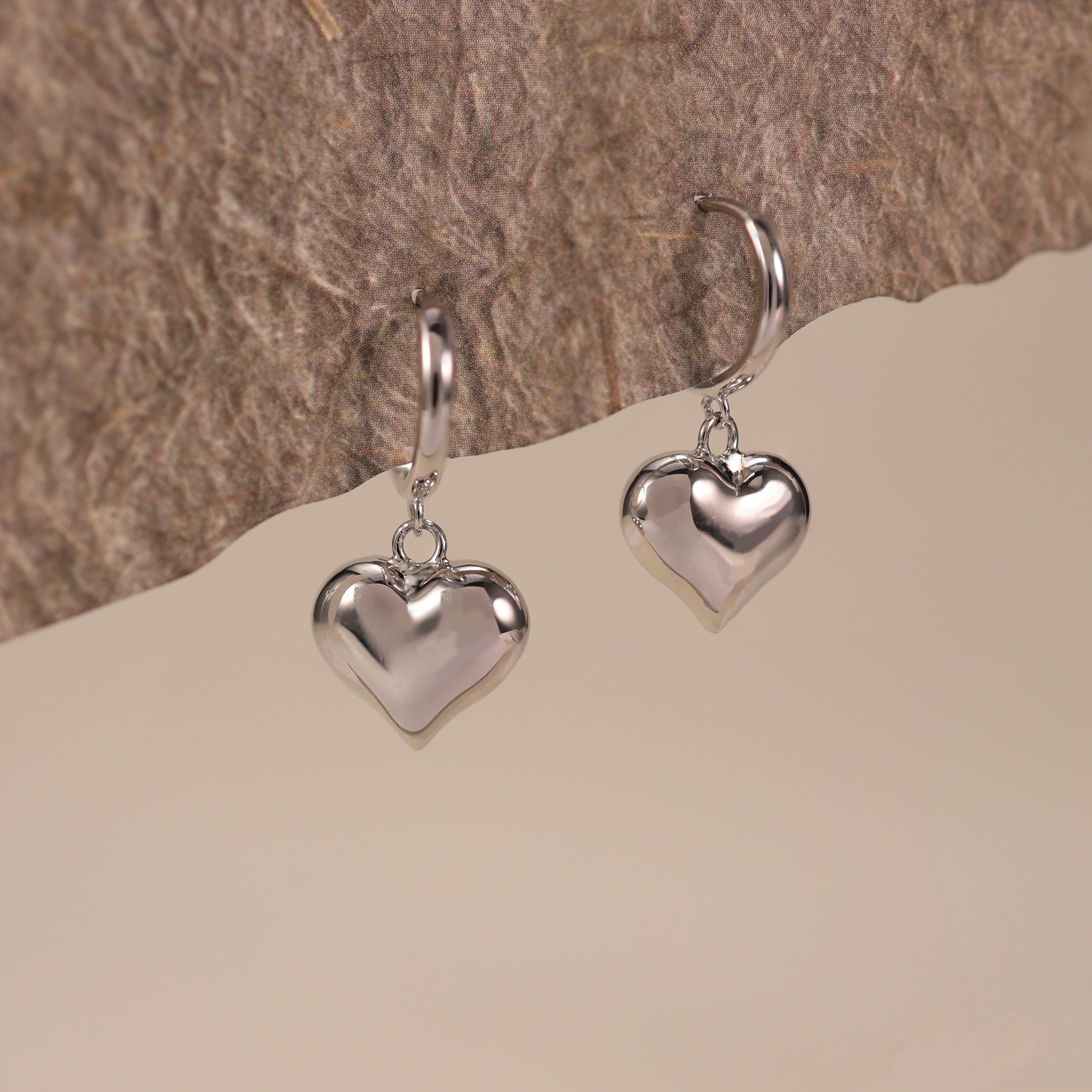 Silvbreeze Lune white gold vermeil heart huggie earrings hanging display, shiny puffy heart charm earrings with polished reflective finish, elegant lifestyle jewelry photography