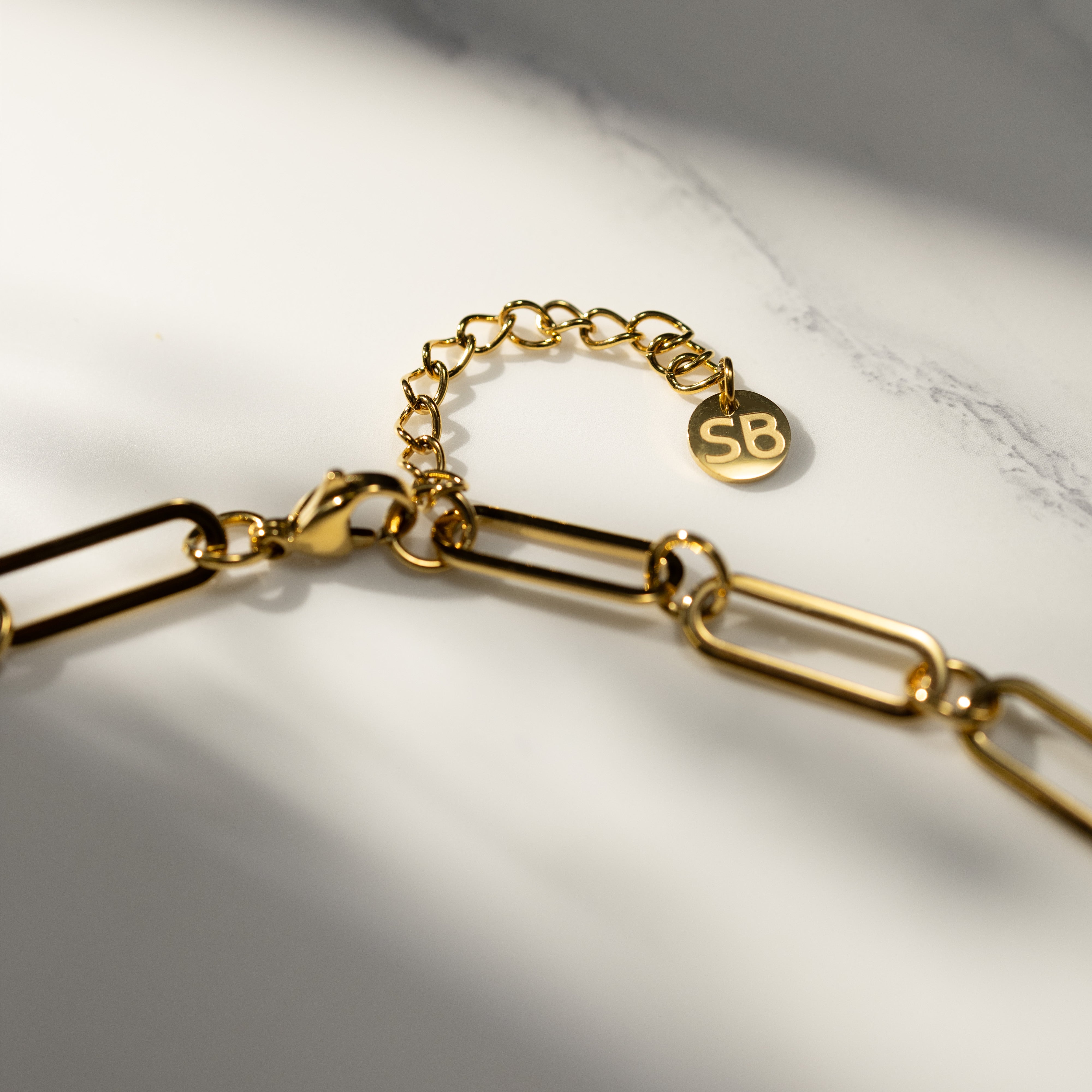 Closeup of the 18K gold MBTI paperclip chain necklace featuring a lobster clasp, adjustable extender chain, and SB tag under soft natural light.