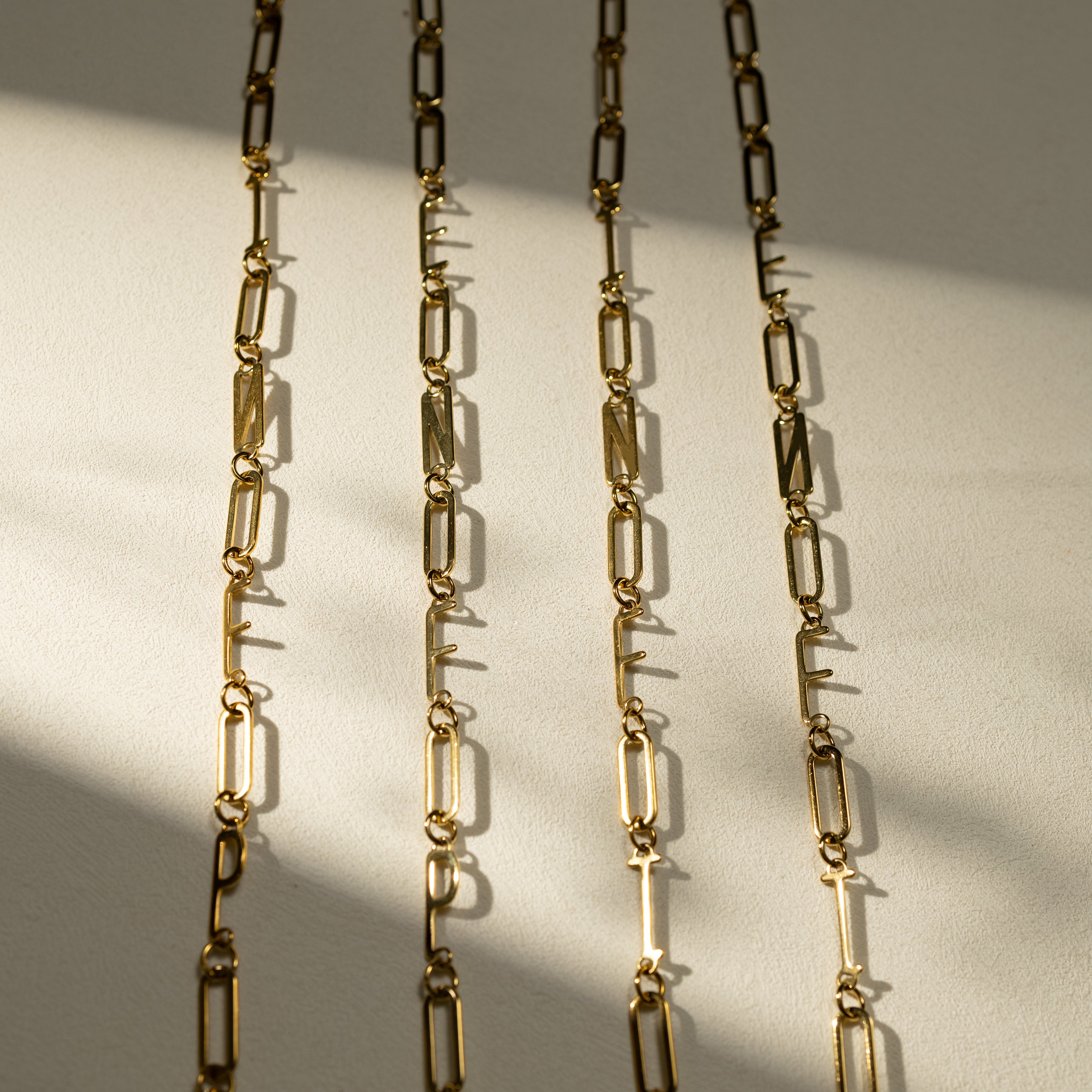 Silvbreeze MBTI 18K gold paperclip chain necklaces featuring INFP, ENFP, INFJ, and ENFJ letters displayed in a vertical lineup under warm natural sunlight on a beige textured surface.