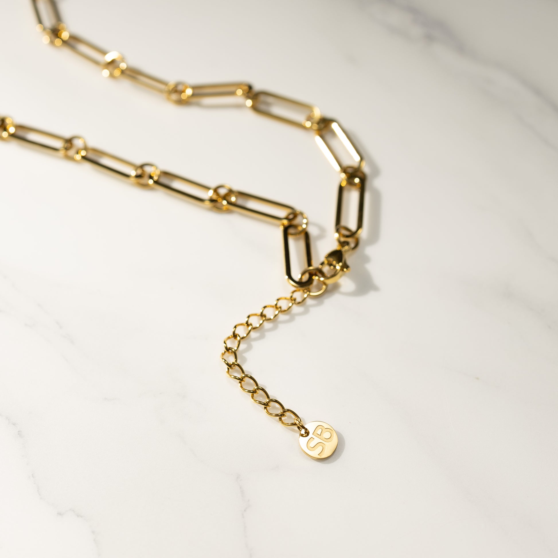 Closeup detail of a gold paperclip MBTI necklace with extension chain and SB tag on a marble background, 18K PVD finish.