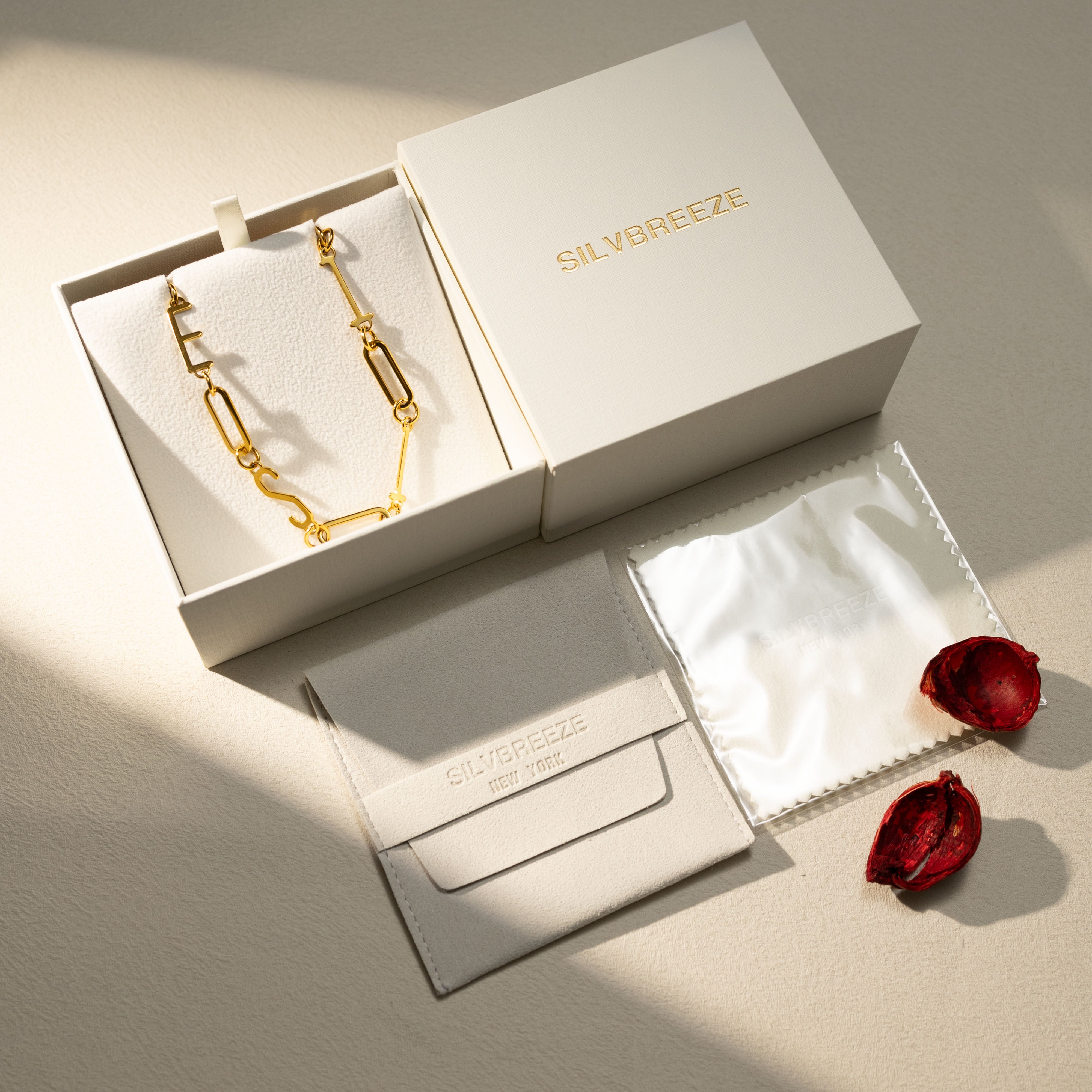 Flatlay of the Silvbreeze MBTI 18K gold-plated ESTJ personality necklace presented in an open gift box with pouch, polishing cloth, and dried red petals under warm natural light.