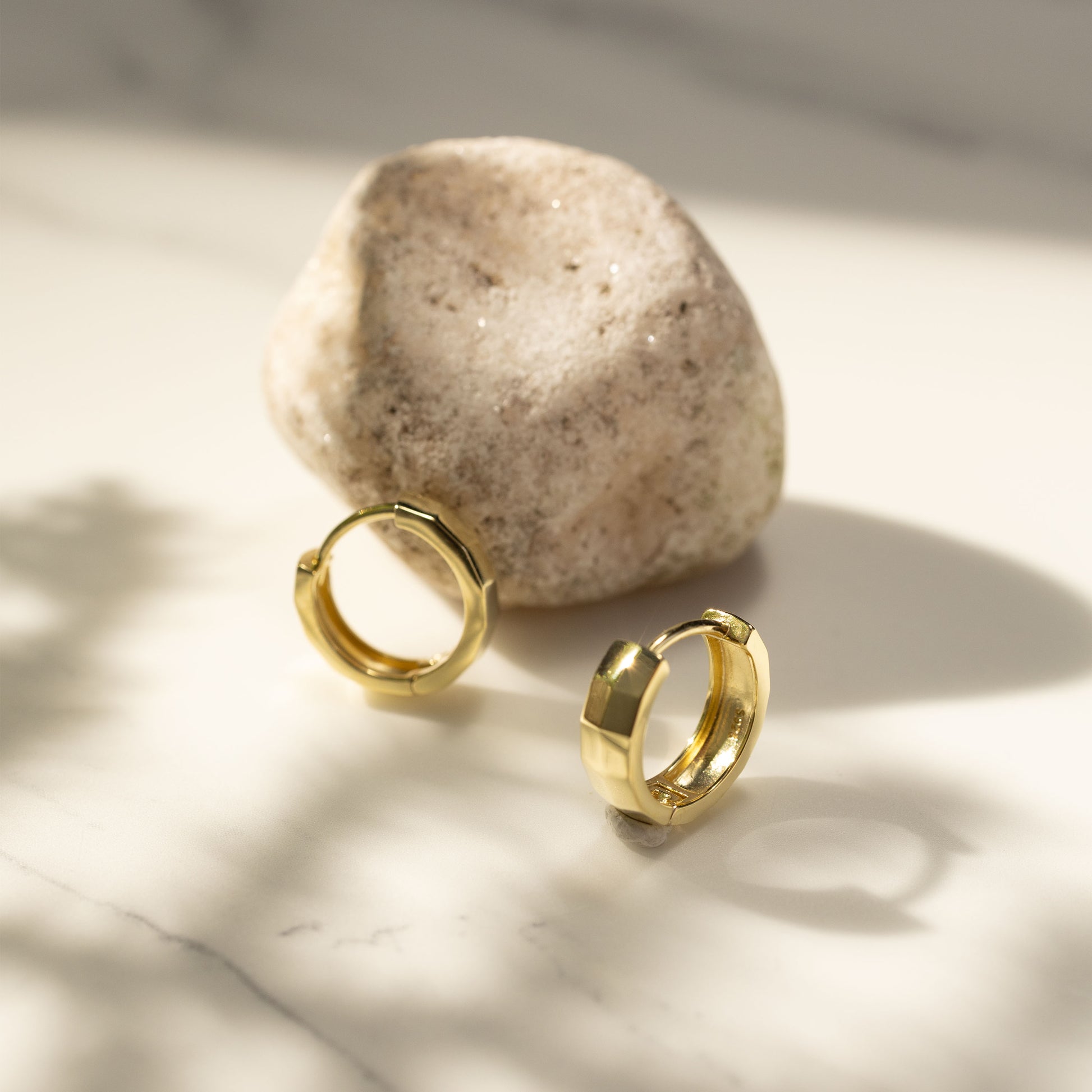 Silvbreeze Mira gold huggie earrings placed on a marble surface beside a natural stone, captured in warm sunlight for a minimal lifestyle jewelry scene.