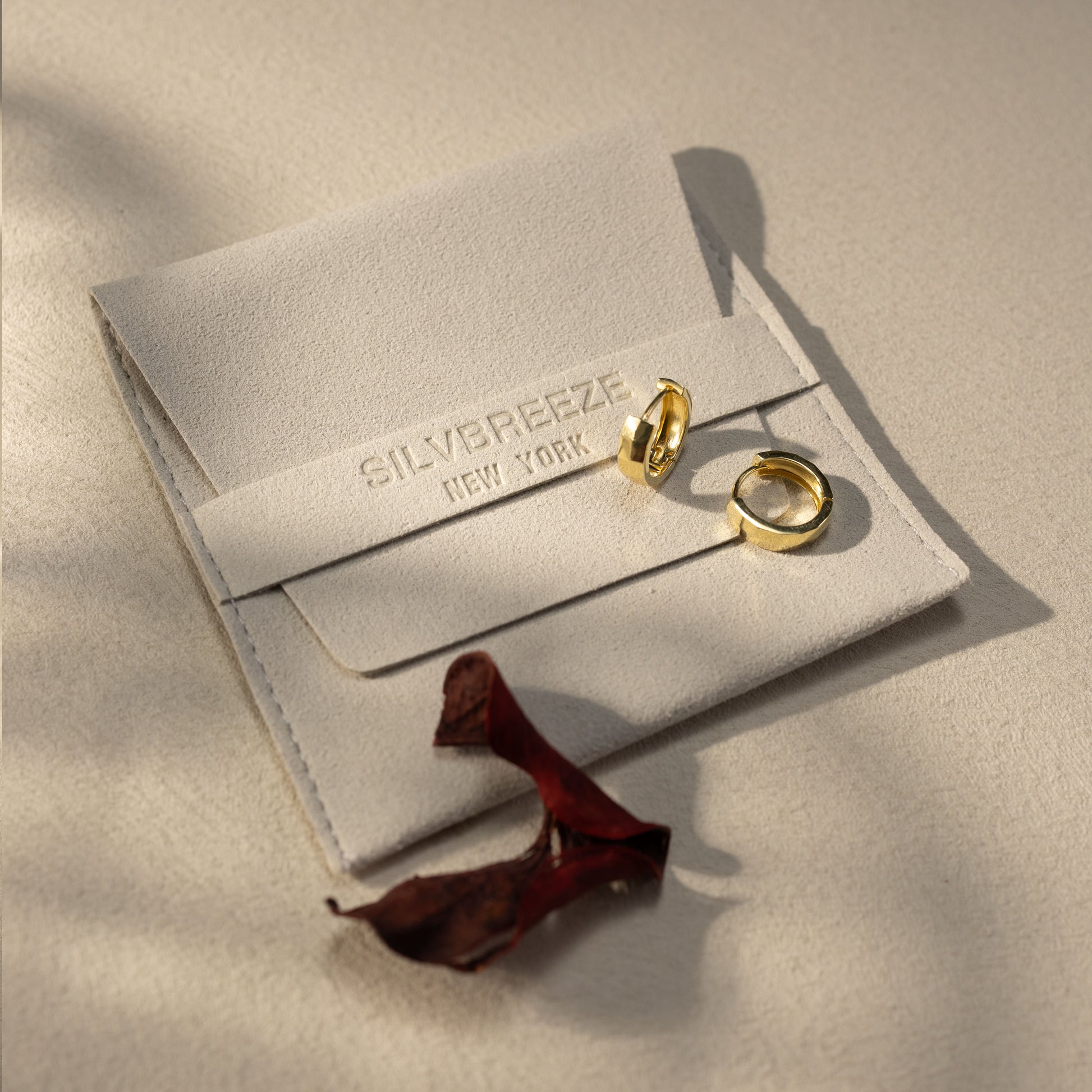 Silvbreeze Mira gold huggie earrings resting on a soft beige suede jewelry pouch, with warm natural sunlight and minimal lifestyle packaging scene.