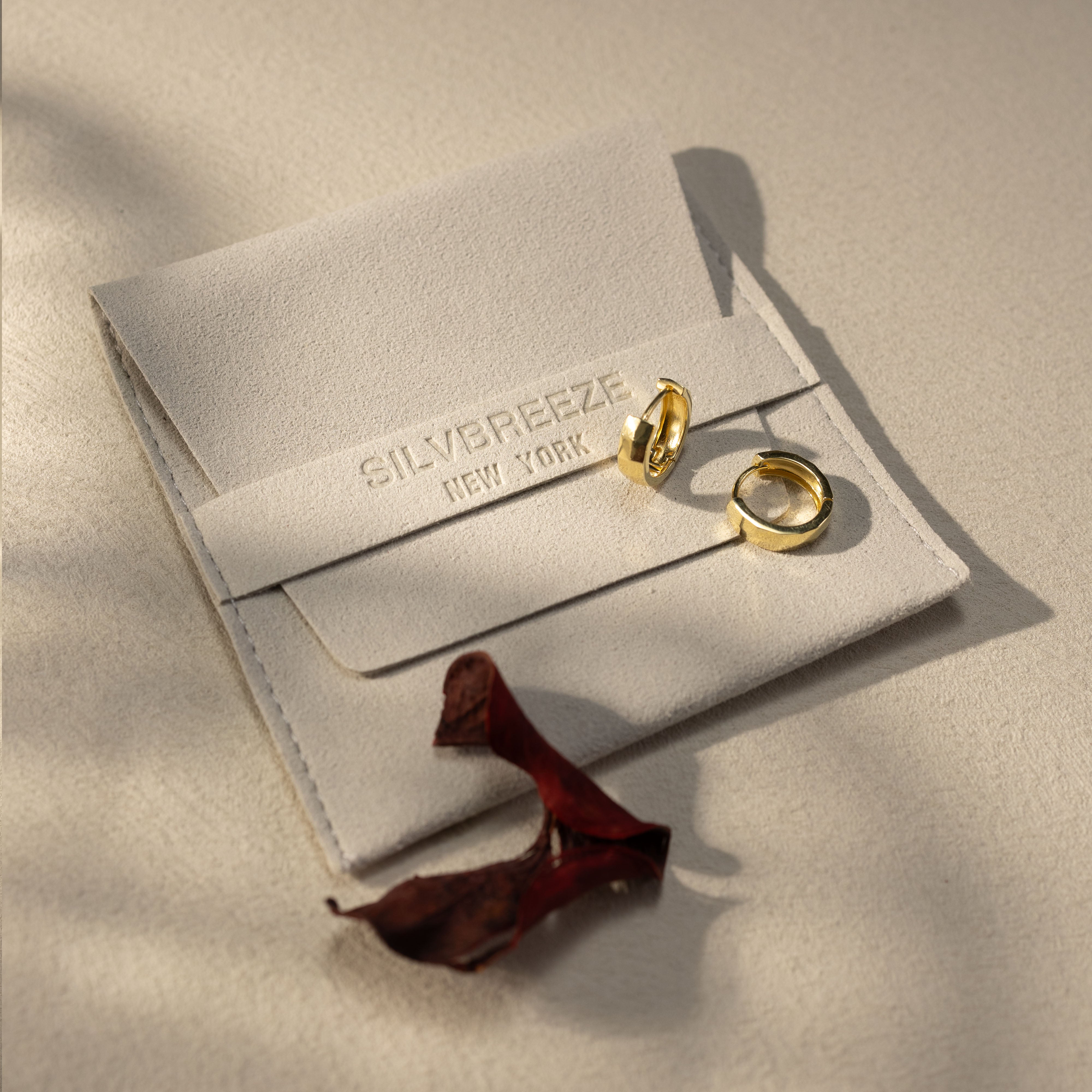 Silvbreeze Mira gold huggie earrings resting on a soft beige suede jewelry pouch, with warm natural sunlight and minimal lifestyle packaging scene.
