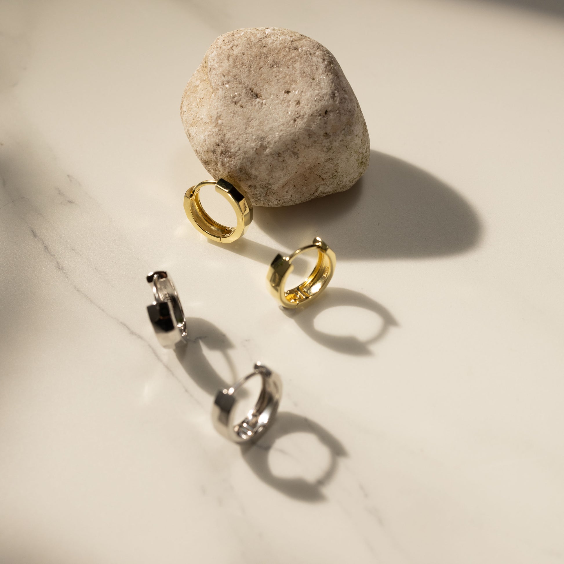 Silvbreeze Mira gold and silver huggie earrings displayed on a marble surface beside a natural stone, captured in warm sunlight for a minimalist lifestyle jewelry scene.