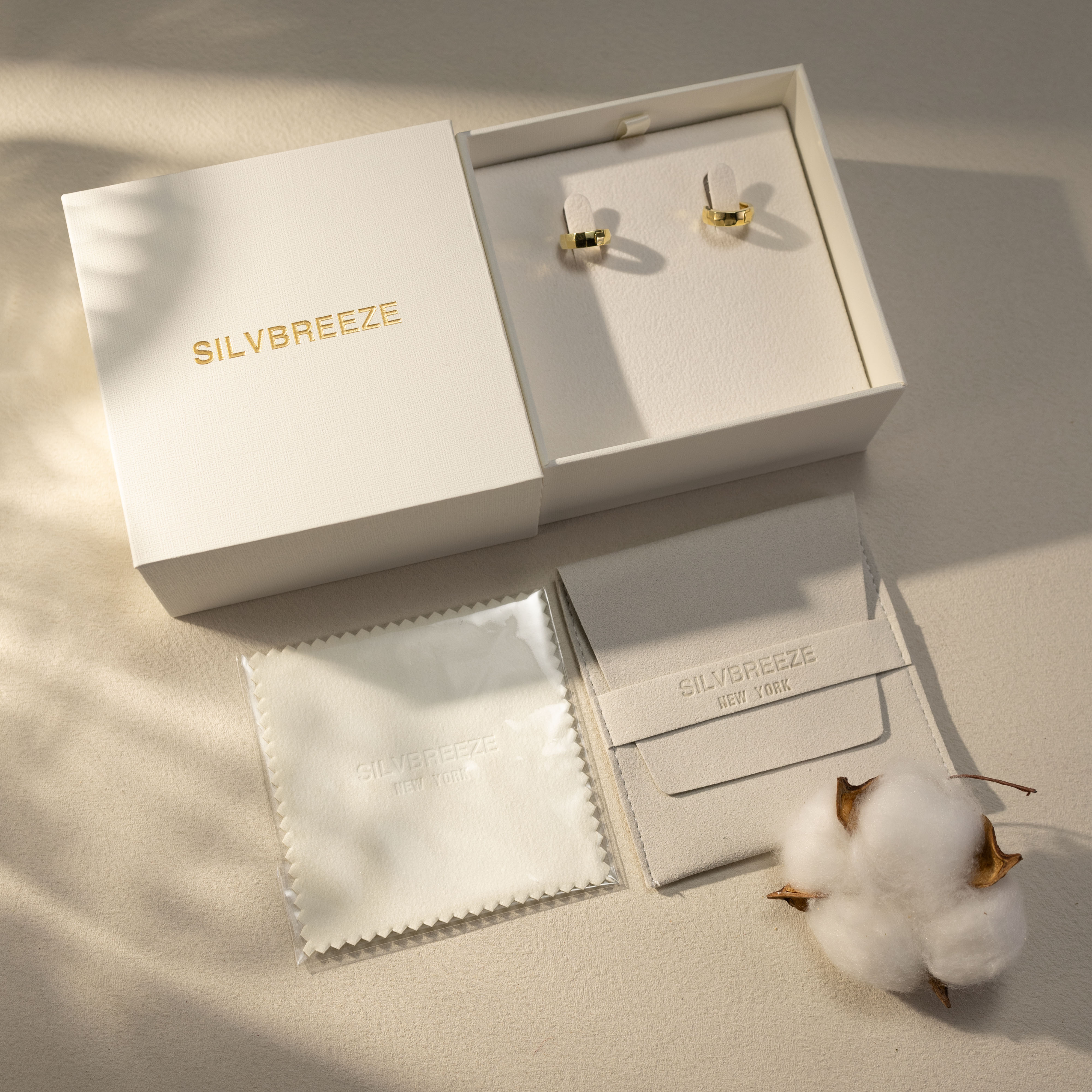Silvbreeze Mira gold huggie earrings presented inside a premium beige jewelry box, displayed with branded suede pouch, polishing cloth, and cotton decor in a soft sunlit packaging scene.