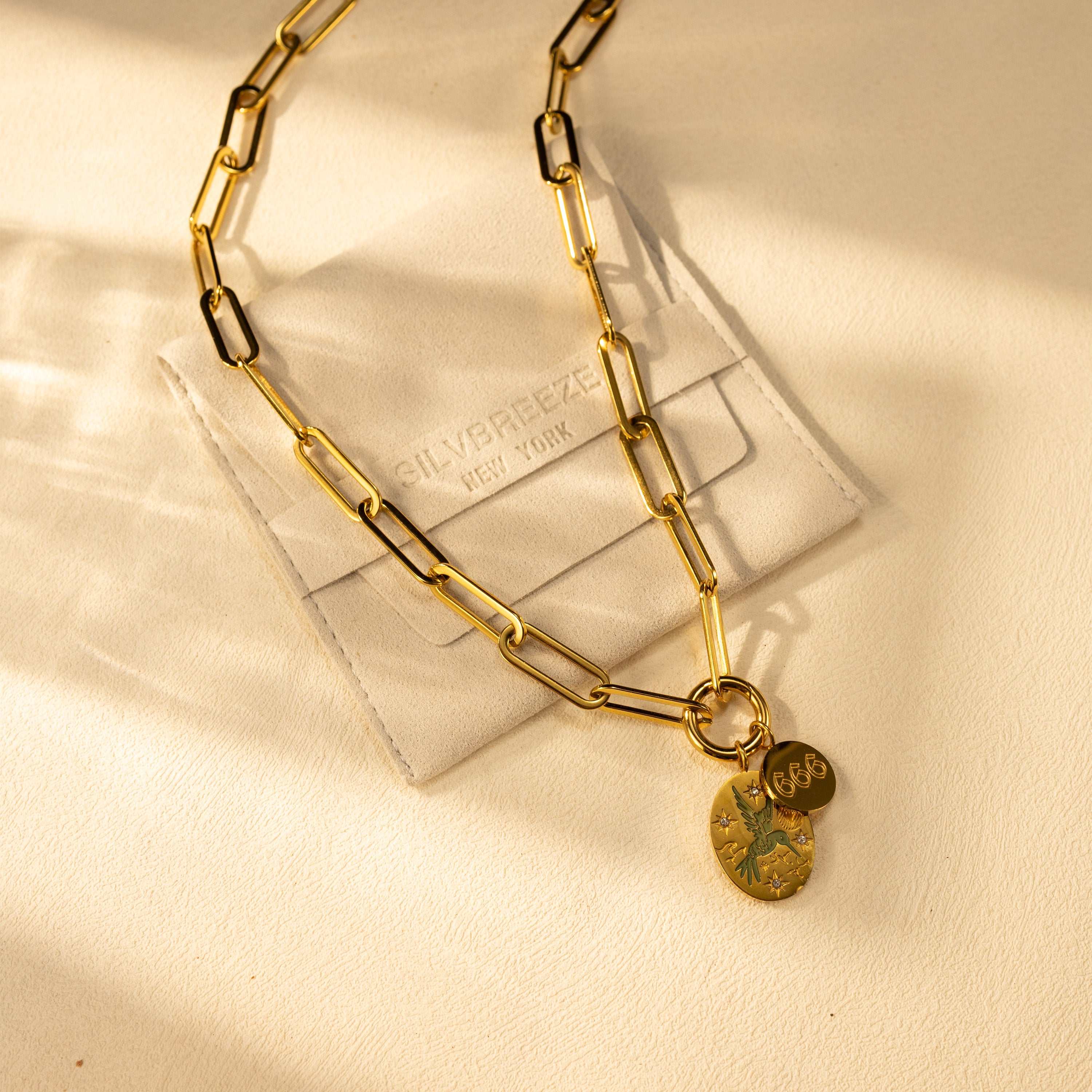 Lifestyle photo of an 18K gold-plated paperclip O-clasp charm holder necklace with a hand-painted green bird pendant and angel number disc, displayed on a SILVBREEZE suede jewelry pouch.