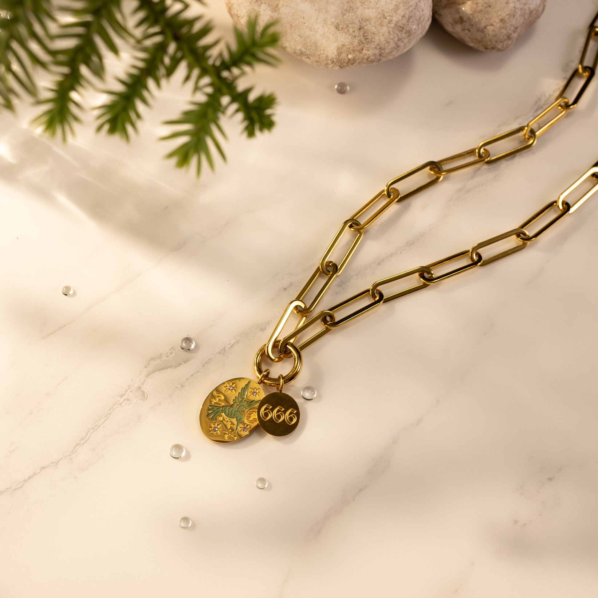18K gold-plated paperclip O-clasp charm holder necklace with a laser-engraved hand-painted green bird pendant and an angel number 666 disc on a marble background.
