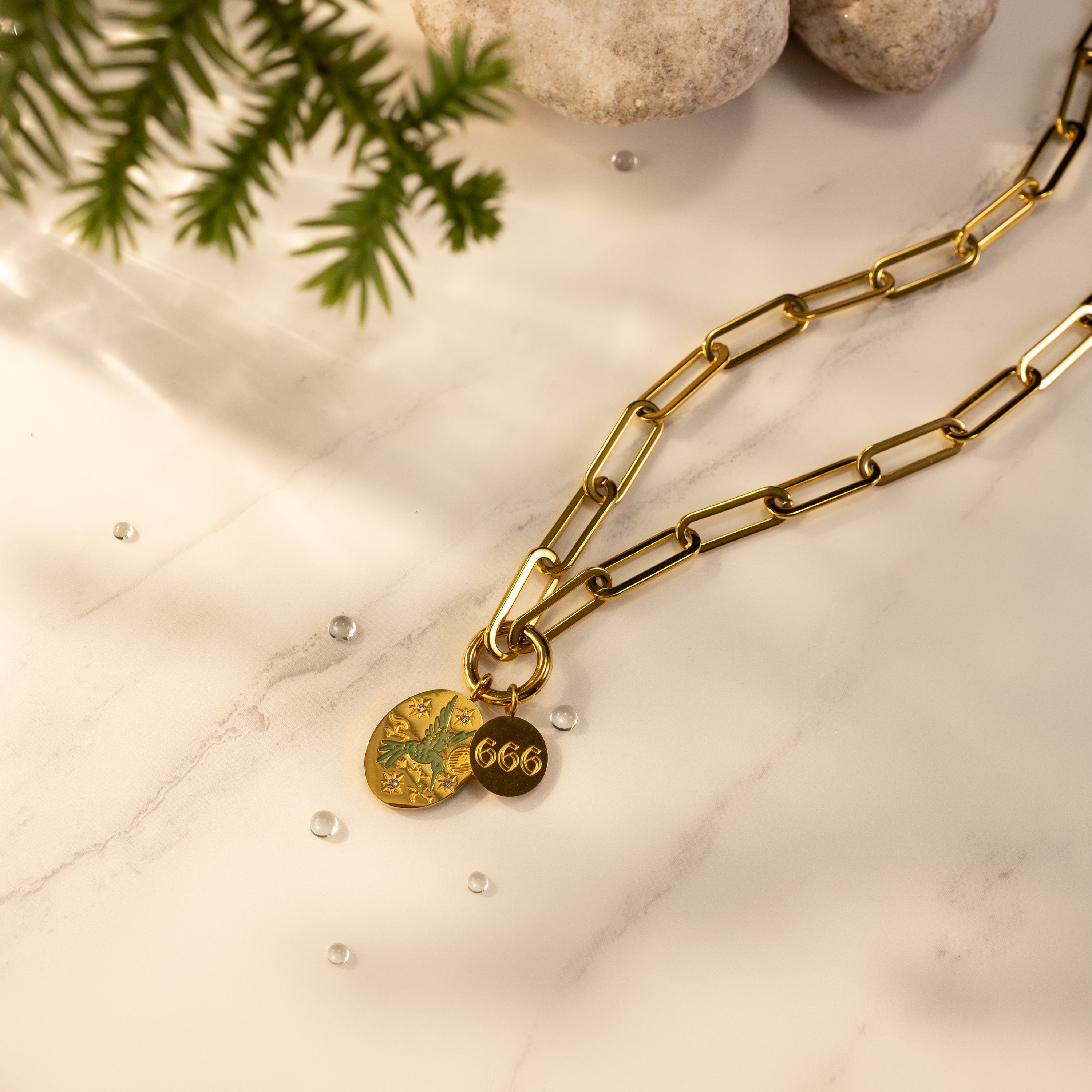 18K gold-plated paperclip O-clasp charm holder necklace with a laser-engraved hand-painted green bird pendant and an angel number 666 disc on a marble background.