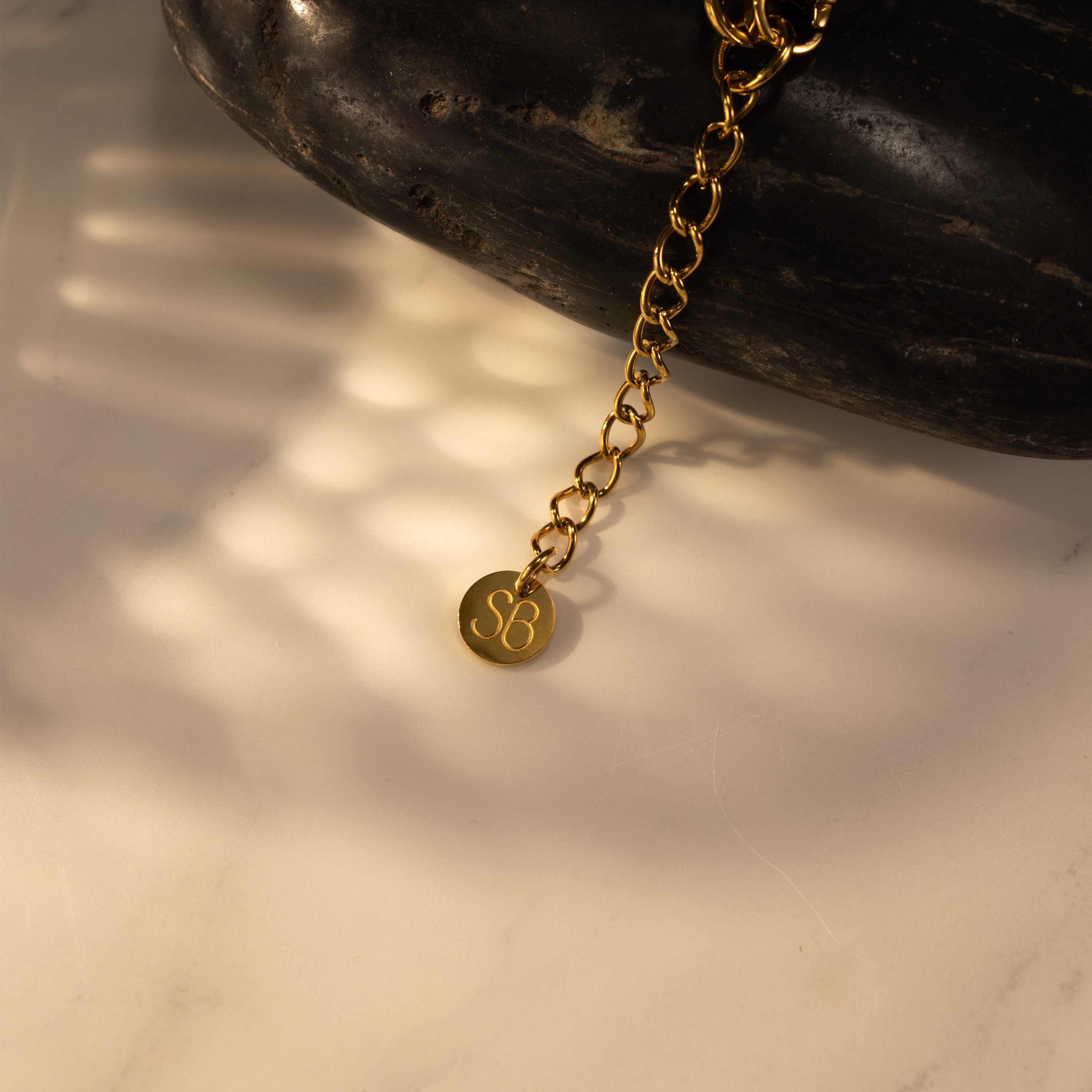 Close-up of the SB logo extender chain tag on an 18K gold-plated SILVBREEZE necklace, showing the adjustable tail chain.