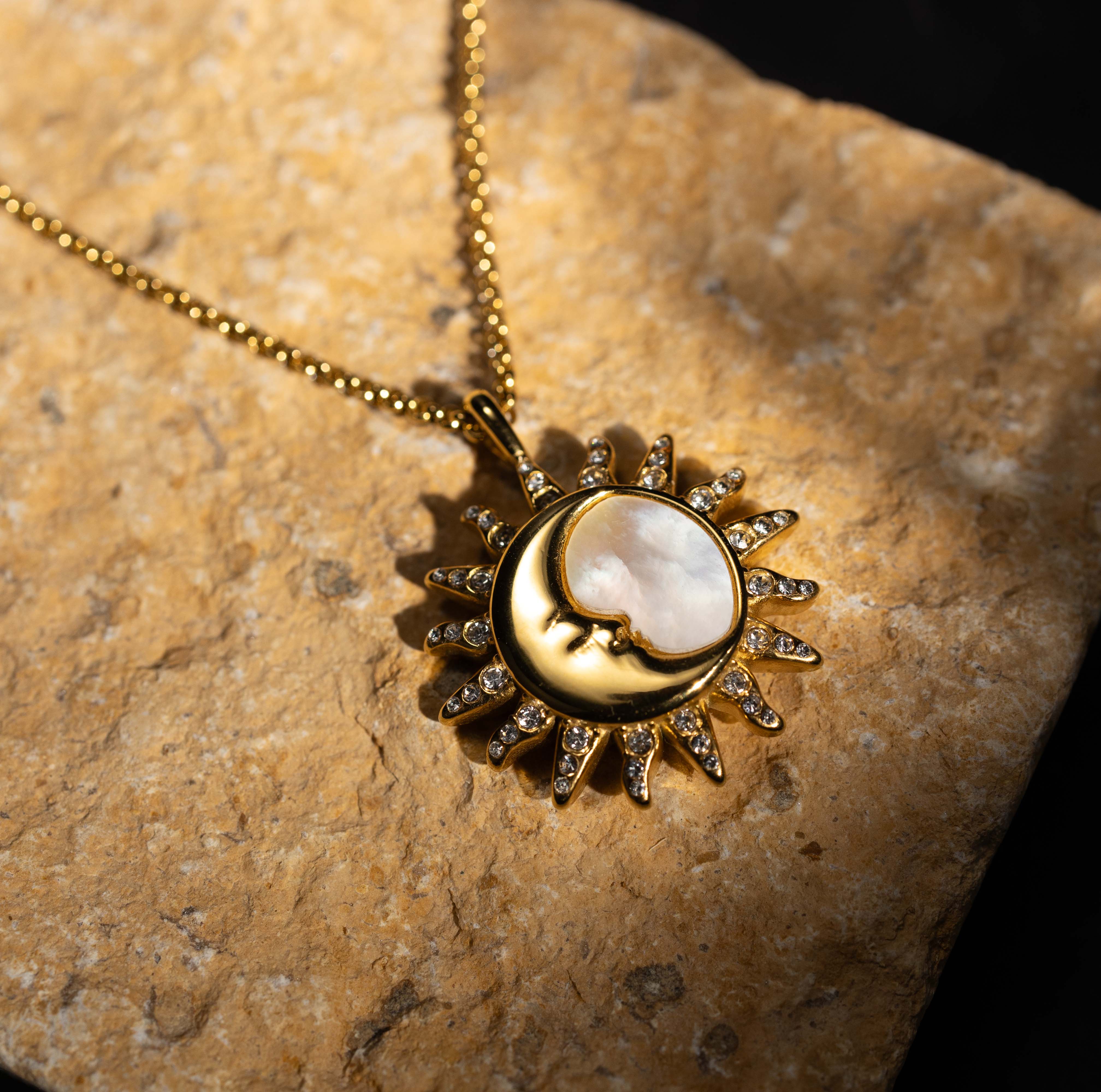 The Silvbreeze "Selene Sol" necklace is captured in a dramatic, angled close-up on a rugged stone surface. The design, a modern celestial talisman, features a gold sun and a serene mother-of-pearl crescent moon with sparkling crystal rays, all brightly illuminated by sharp, natural sunlight.