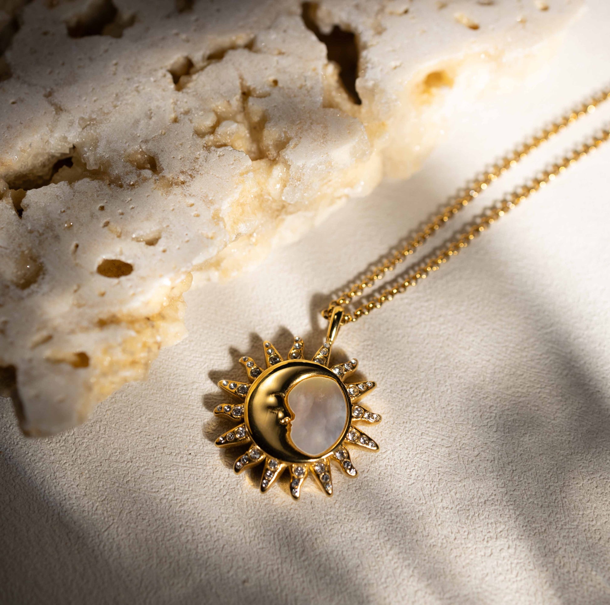 A beautifully composed shot of the Silvbreeze "Selene Sol" necklace, featuring a gold sunburst and mother-of-pearl moon face resting on a light-colored, porous rock. The soft sunlight highlights the intricate crystal pavé and the iridescent texture of the shell inlay.