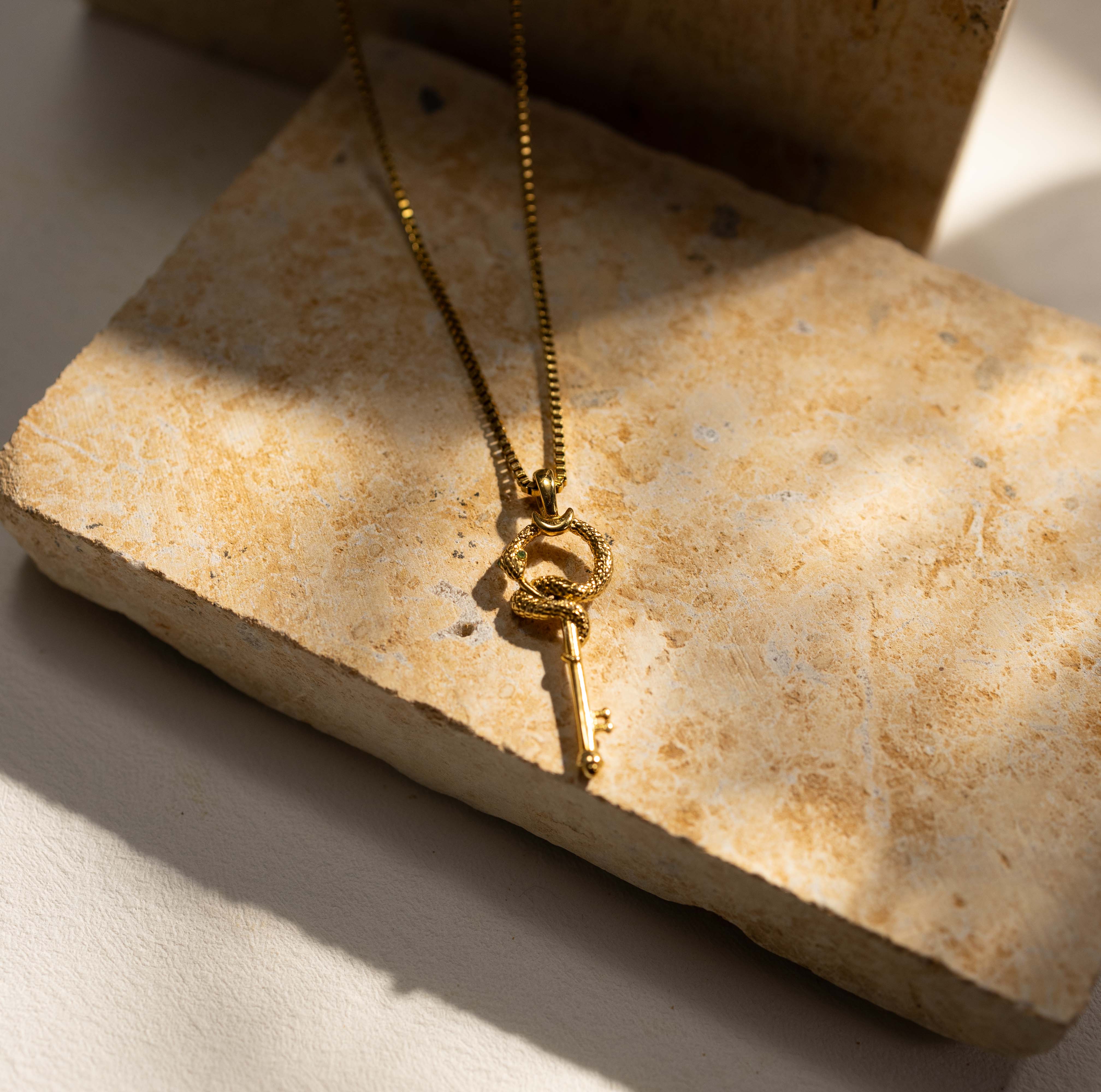 Silvbreeze gold serpent key necklace hanging against natural stone, highlighting the circular serpent form and key pendant