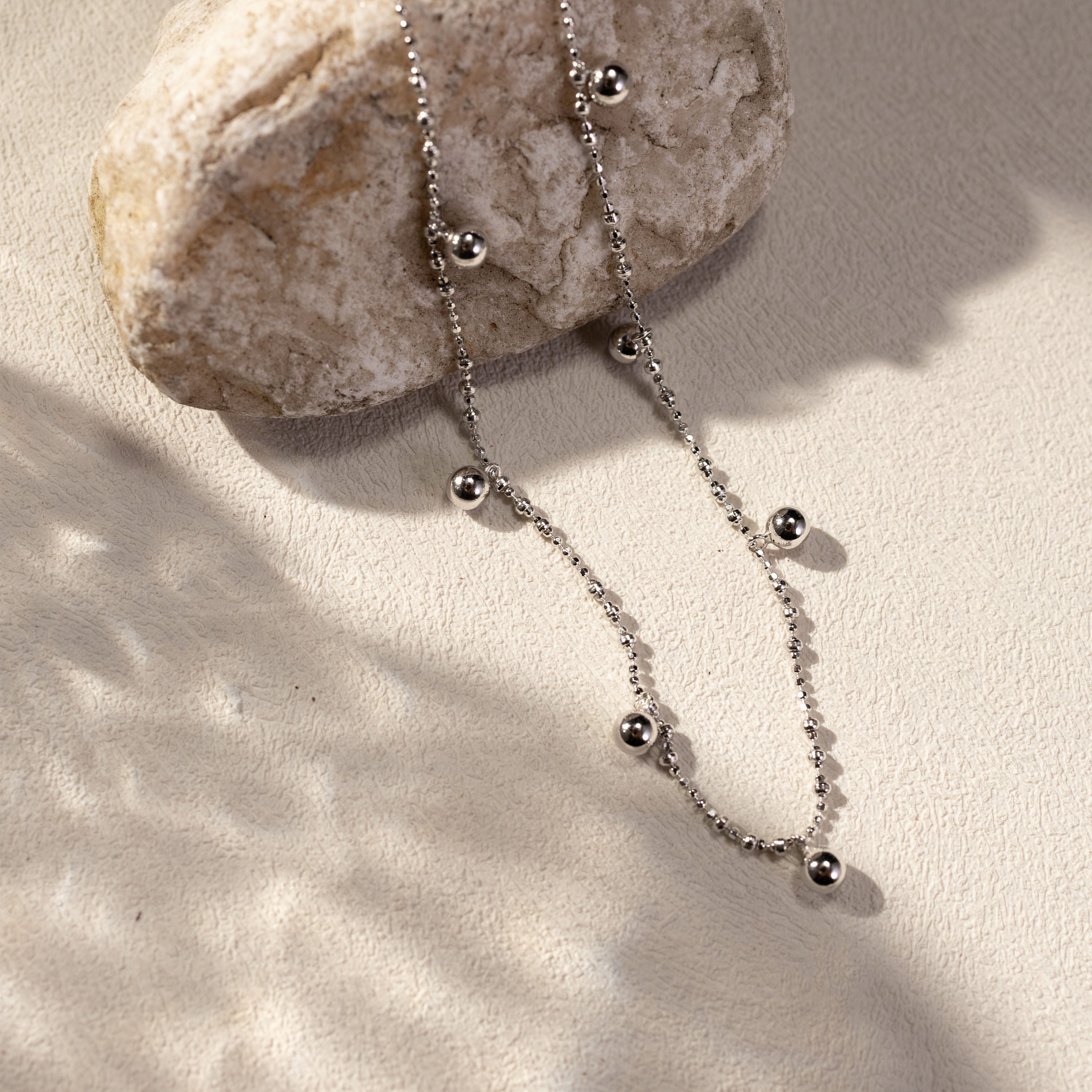 Silver beaded necklace styled on a stone surface with soft warm lighting and delicate shadows.