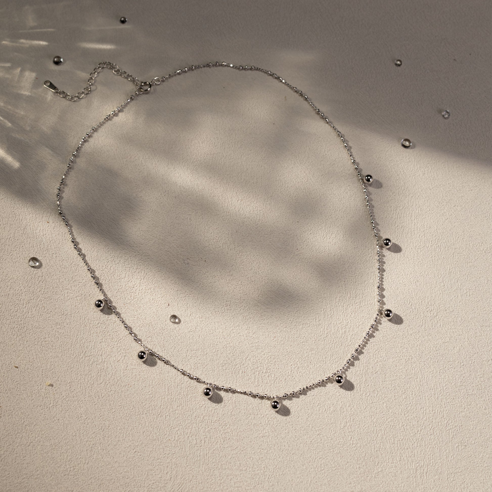 Silver beaded necklace styled flat on a soft textured surface with scattered beads and warm natural shadows