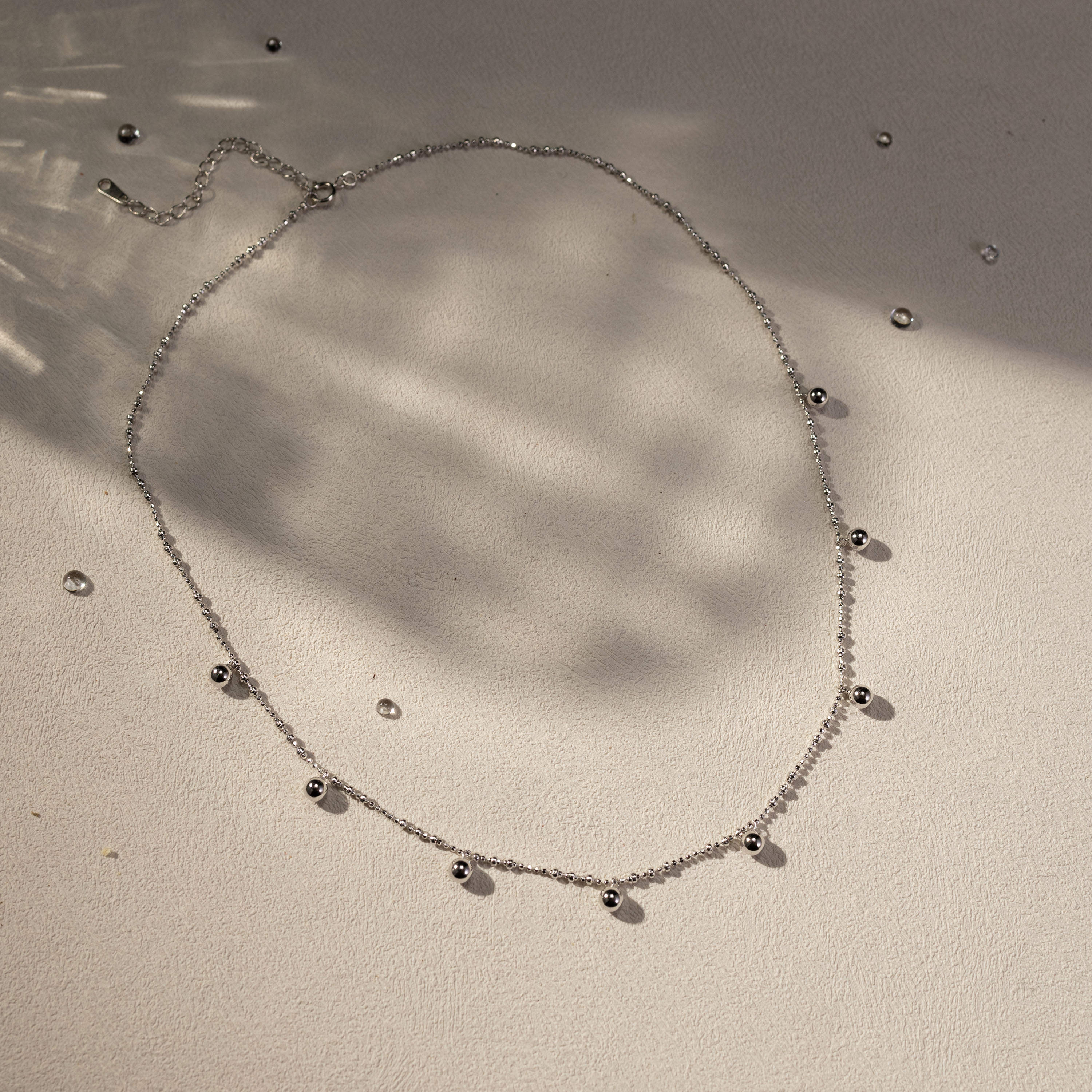 Silver beaded necklace styled flat on a soft textured surface with scattered beads and warm natural shadows