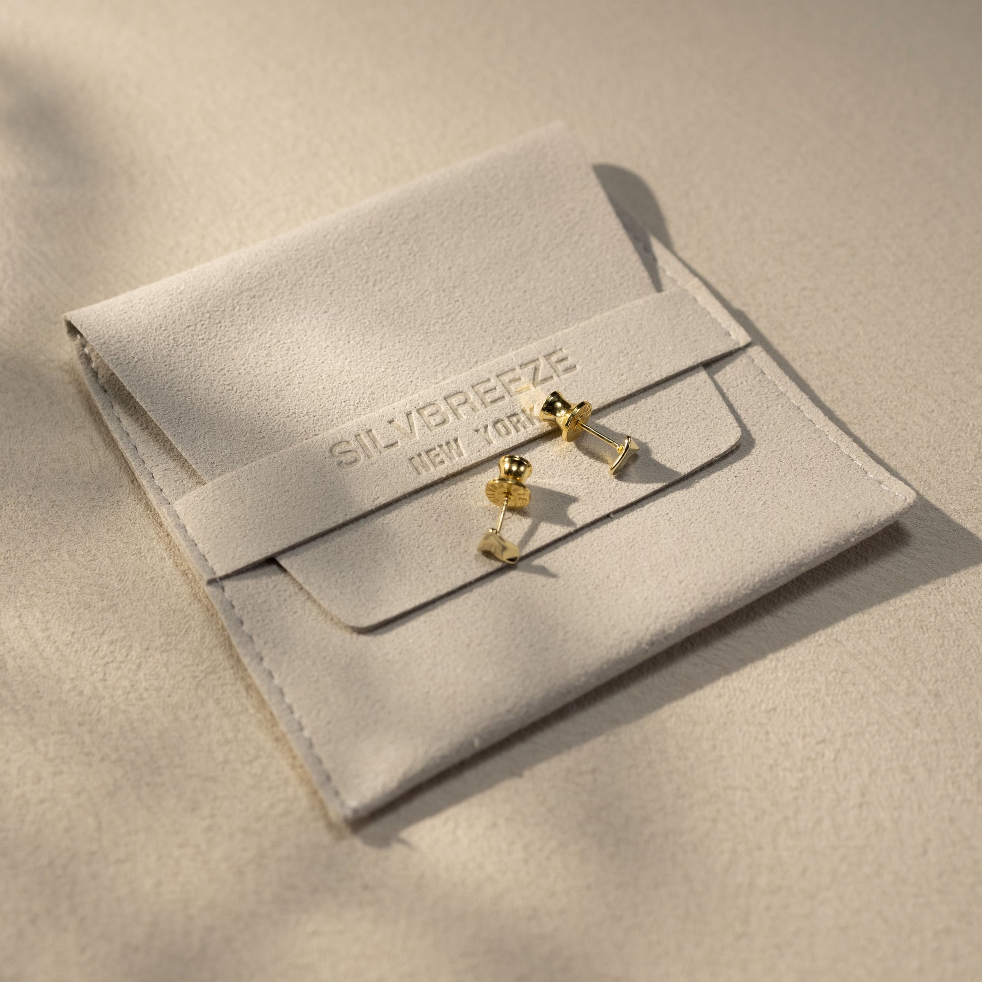 Tria 14K gold vermeil triangle stud earrings displayed on a soft suede Silvbreeze jewelry pouch, captured in warm natural light to highlight premium packaging and elevated lifestyle presentation