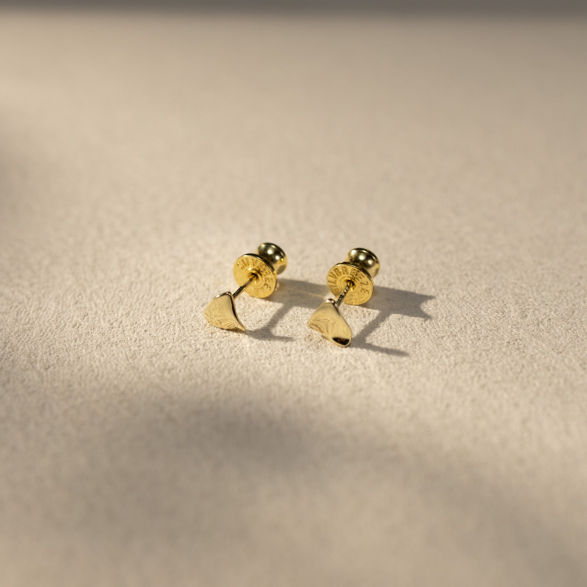 Tria 14K gold vermeil triangle stud earrings with screw backs resting on a soft textured surface, captured in warm natural light with subtle shadows for an elevated lifestyle jewelry look